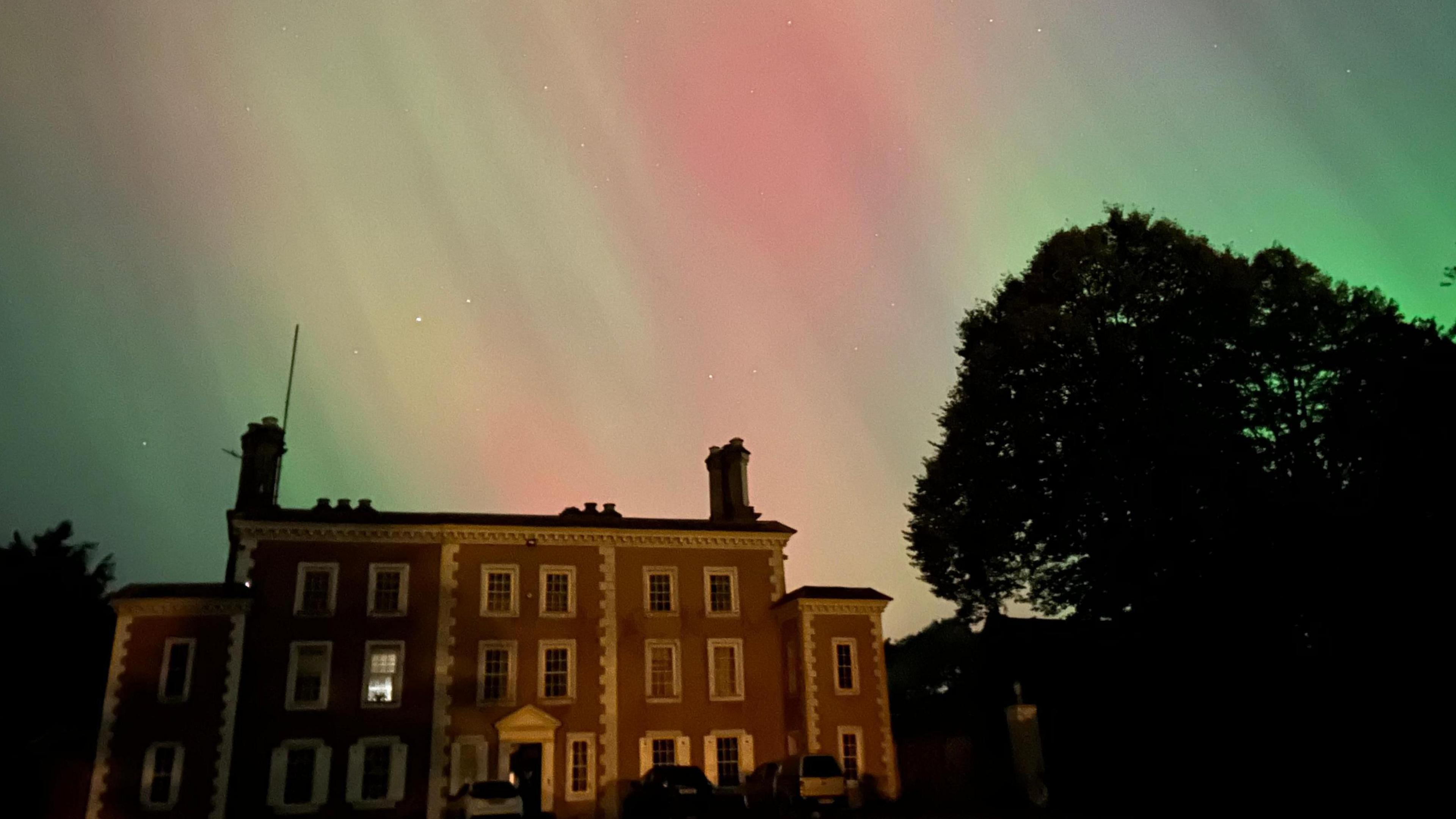 Red and green streams of light are visible in the clear night sky surrounded by stars.  A brown house with white windows sits below surrounded by trees.