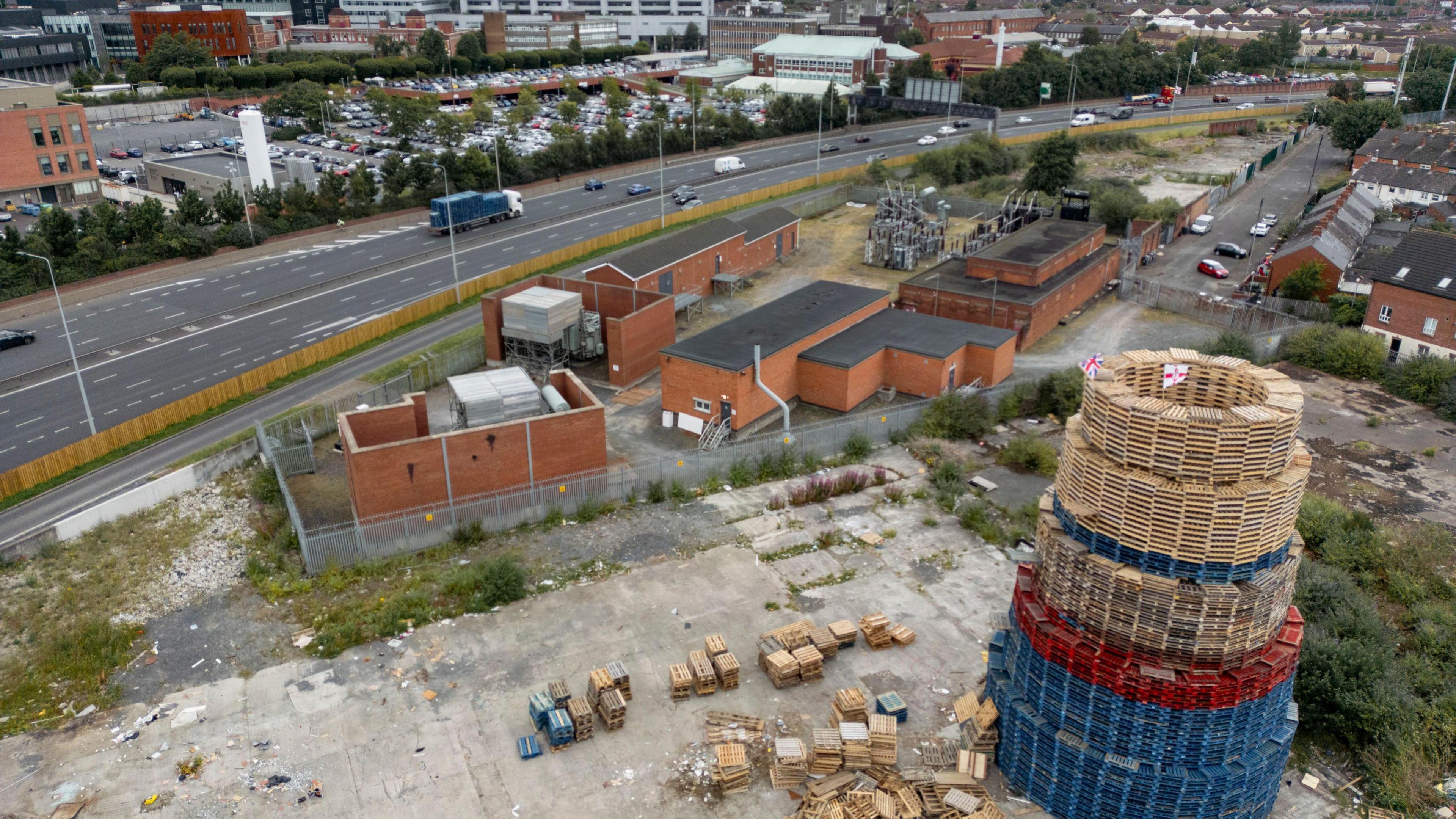 A drone shot of the bonfire, made up of wooden pallets which are blue at the bottom of the structure, red in the middle and plain wood on top. A few metres away towards the top of the image are a series of red brick buildings making up the electricity substation. Beyond this is the Westlink - a dual-carriageway A road.