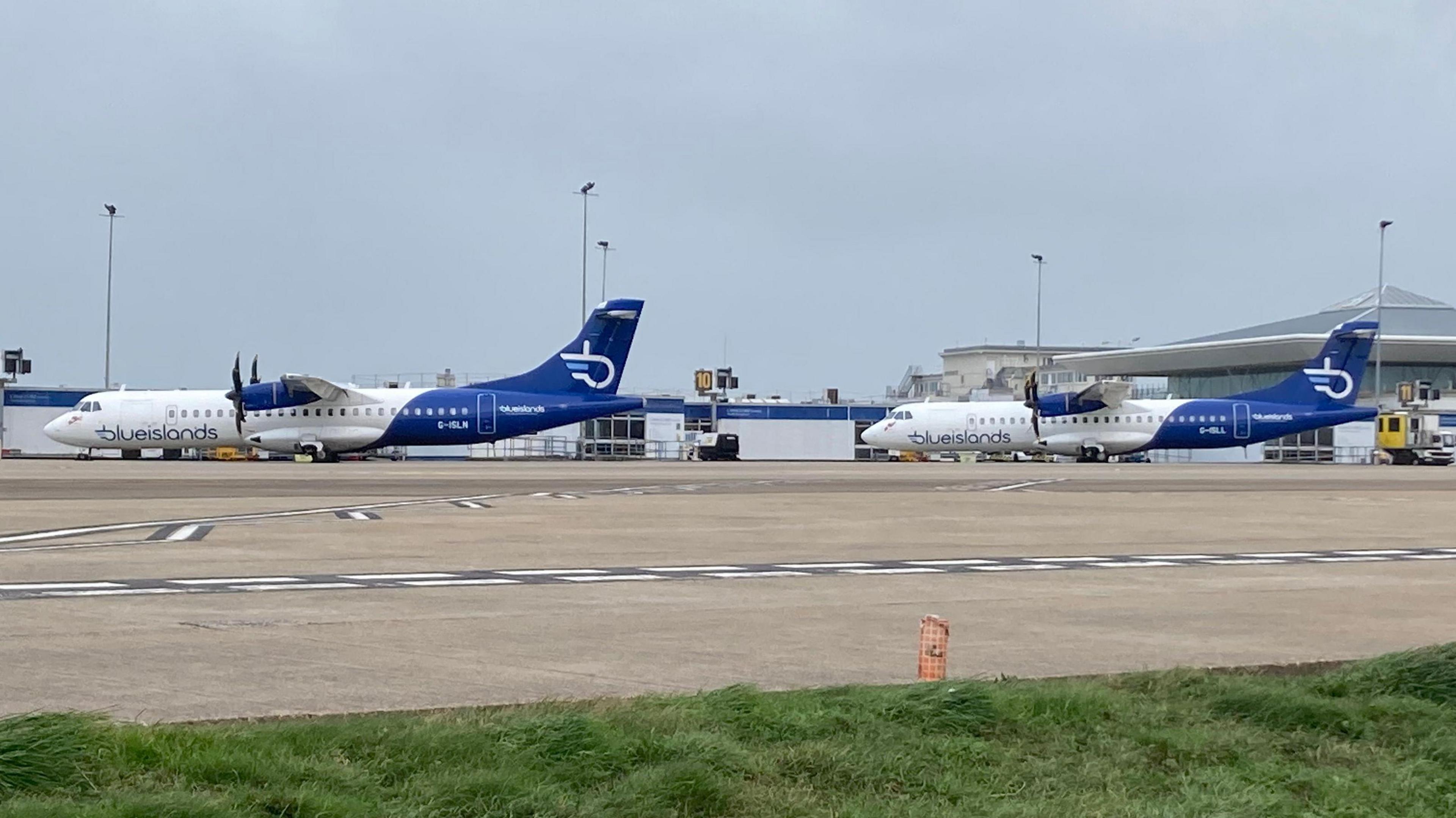 Two Blue Islands planes parked up on the tarmac at an airport. The terminal building is in the background of the shot.