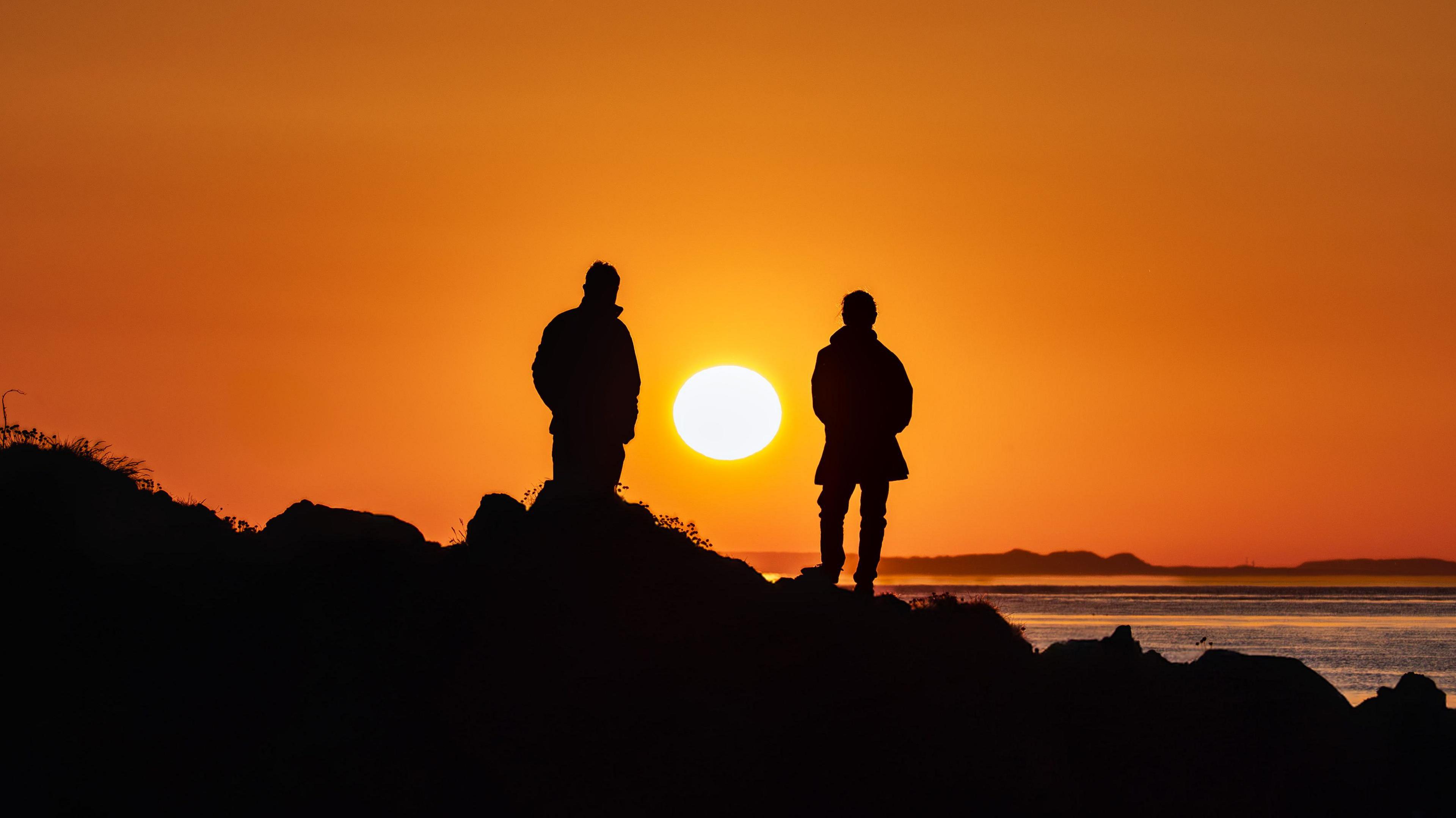 Two silhouetted figures stand on rocks against a vibrant sunset, casting a warm glow over the serene ocean.