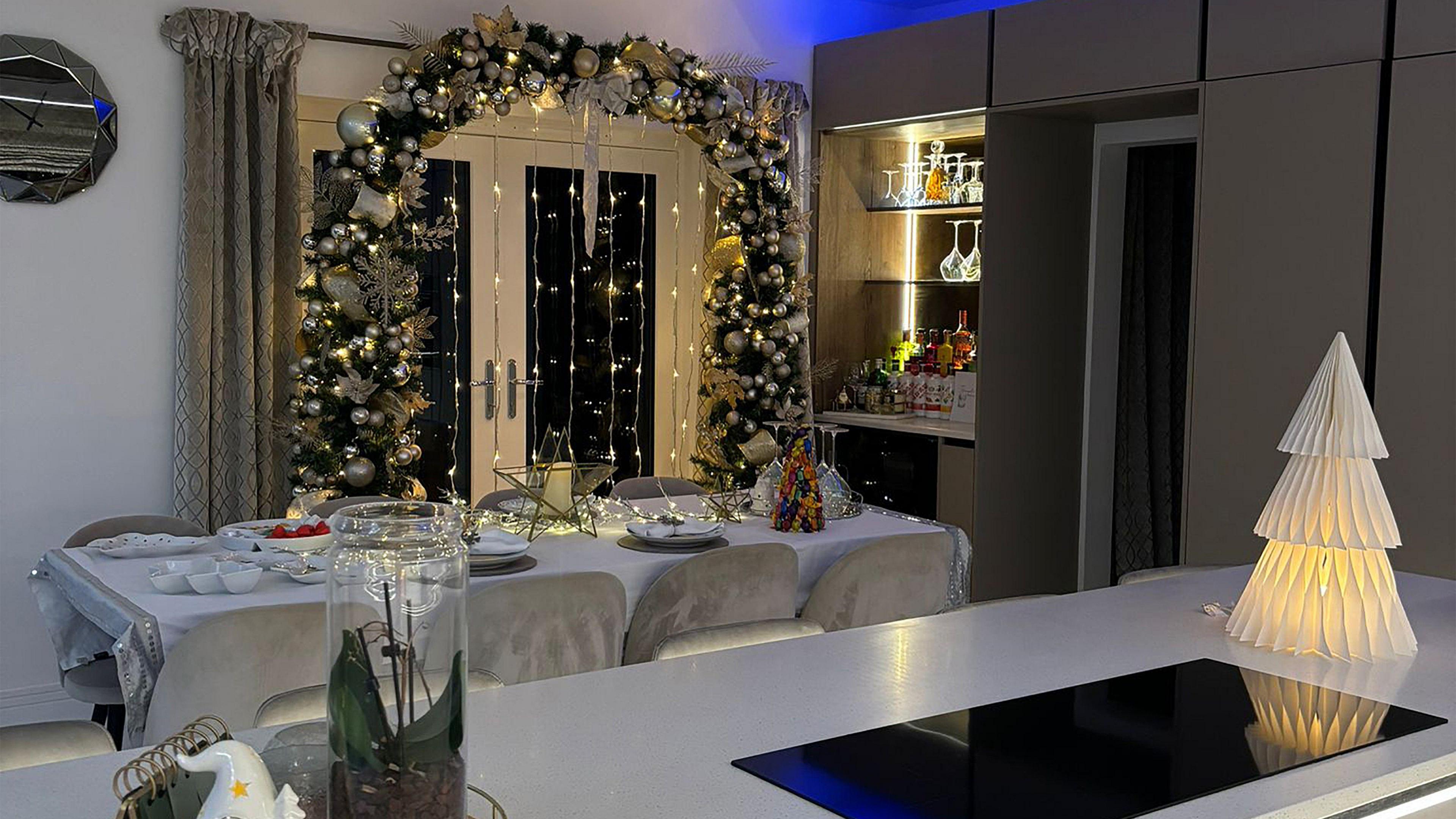 A Christmas themed kitchen. A large archway made of foliage, baubles and warm yellow Christmas lights borders the patio doors. A dining table, covered in fairy lights and a star, looks set for Christmas dinner.