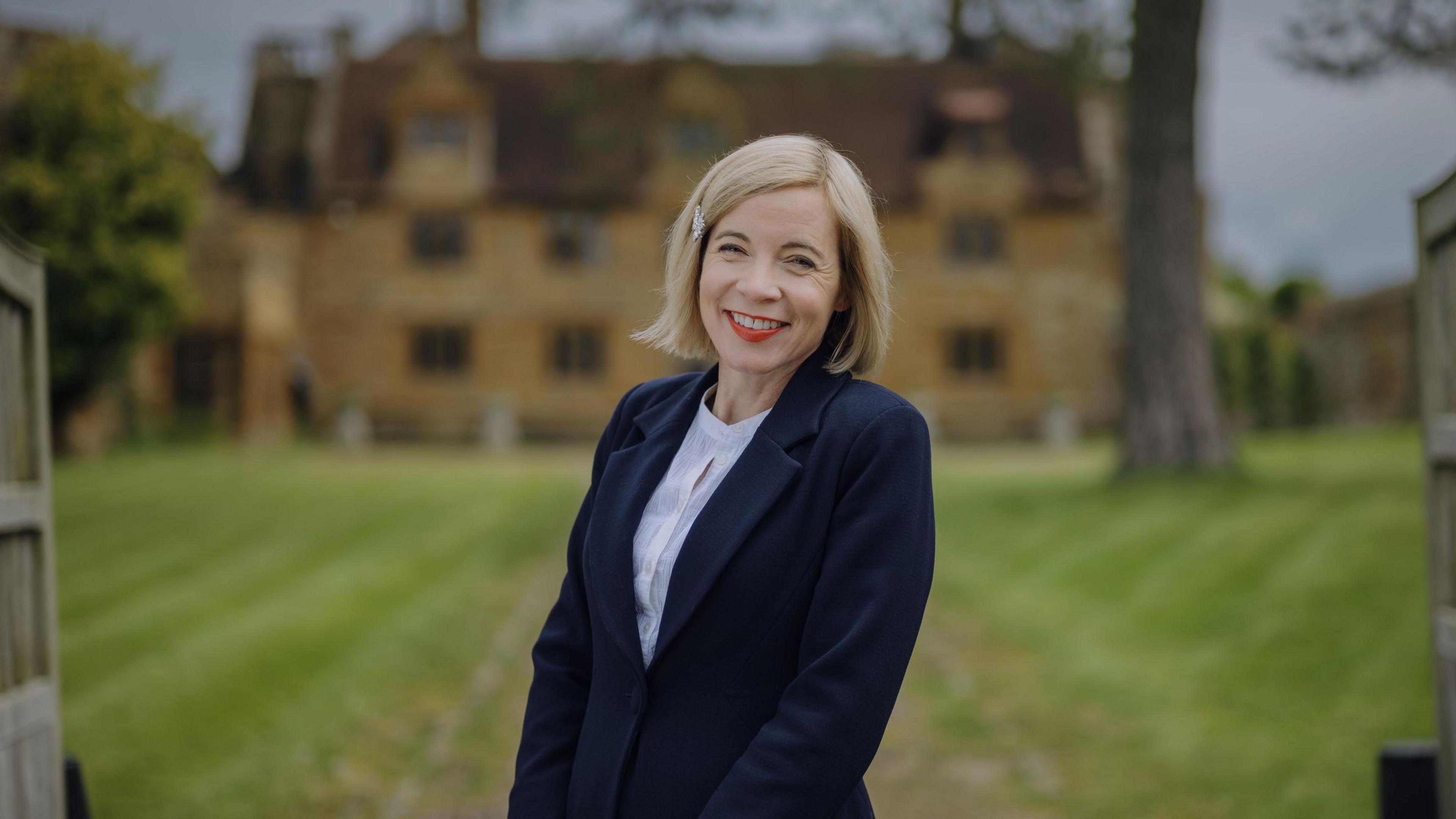 Lucy Worsley standing outside Ashby Manor, St Ledgers. She is wearing a dark coat, light shirt,  is smiling and looking at the camera. She is standing in front of a historic house, with bricks and a large number of windows, the house behind is blurred. Lucy has blond chin length hair, with a side parting, and a clip to the left. 