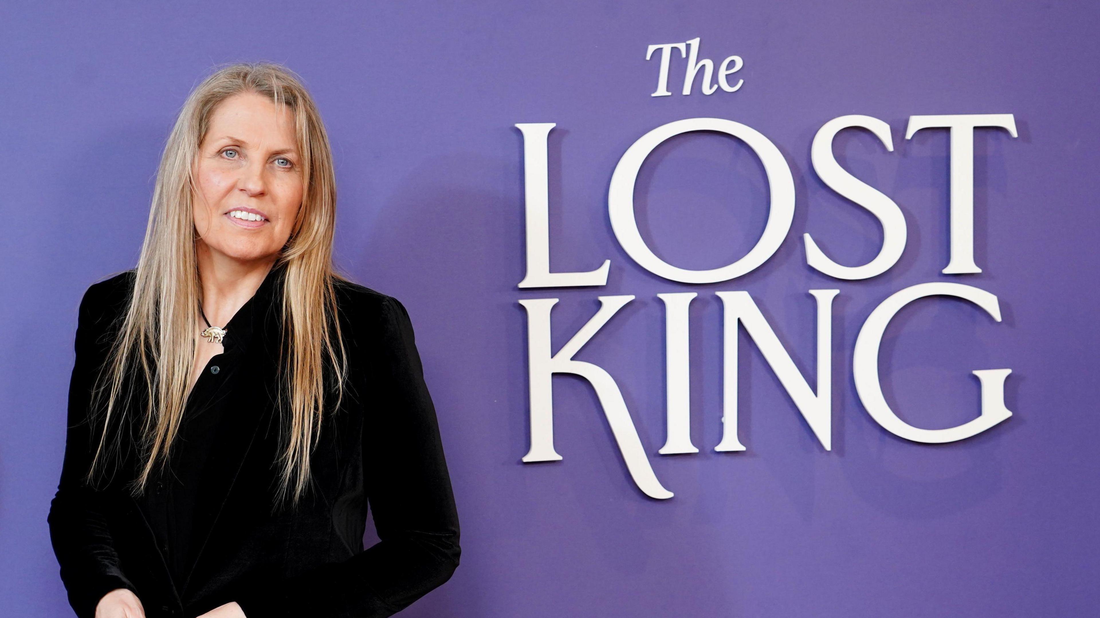 A woman in black standing by a logo saying The Lost King on a purple background