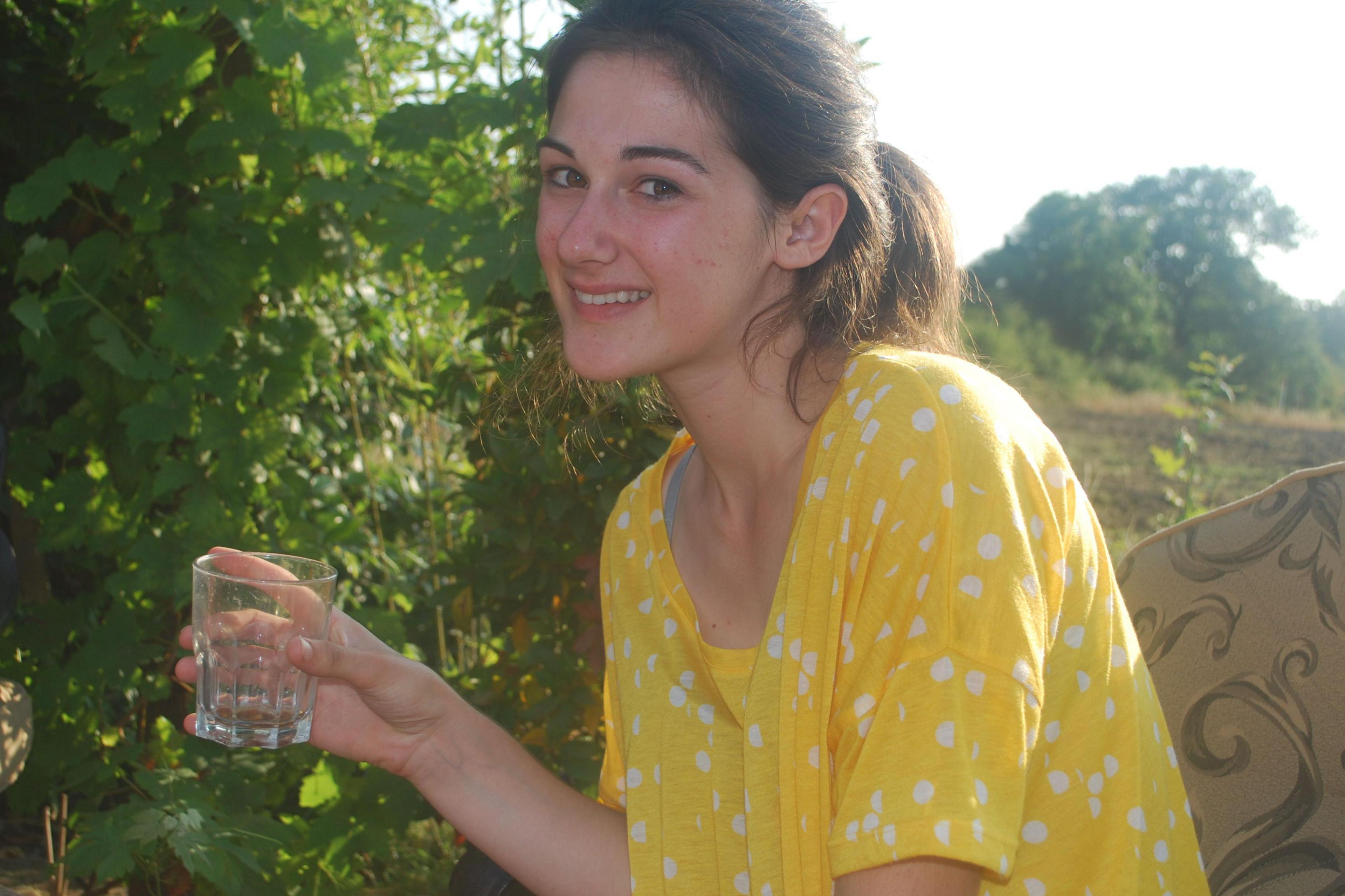 A teenage Amanda Cole holds an empty glass while looking slightly pensively at the camera. She is outside in a yellow polka dot dress in an outdoors location. The sun is shining from behind her.