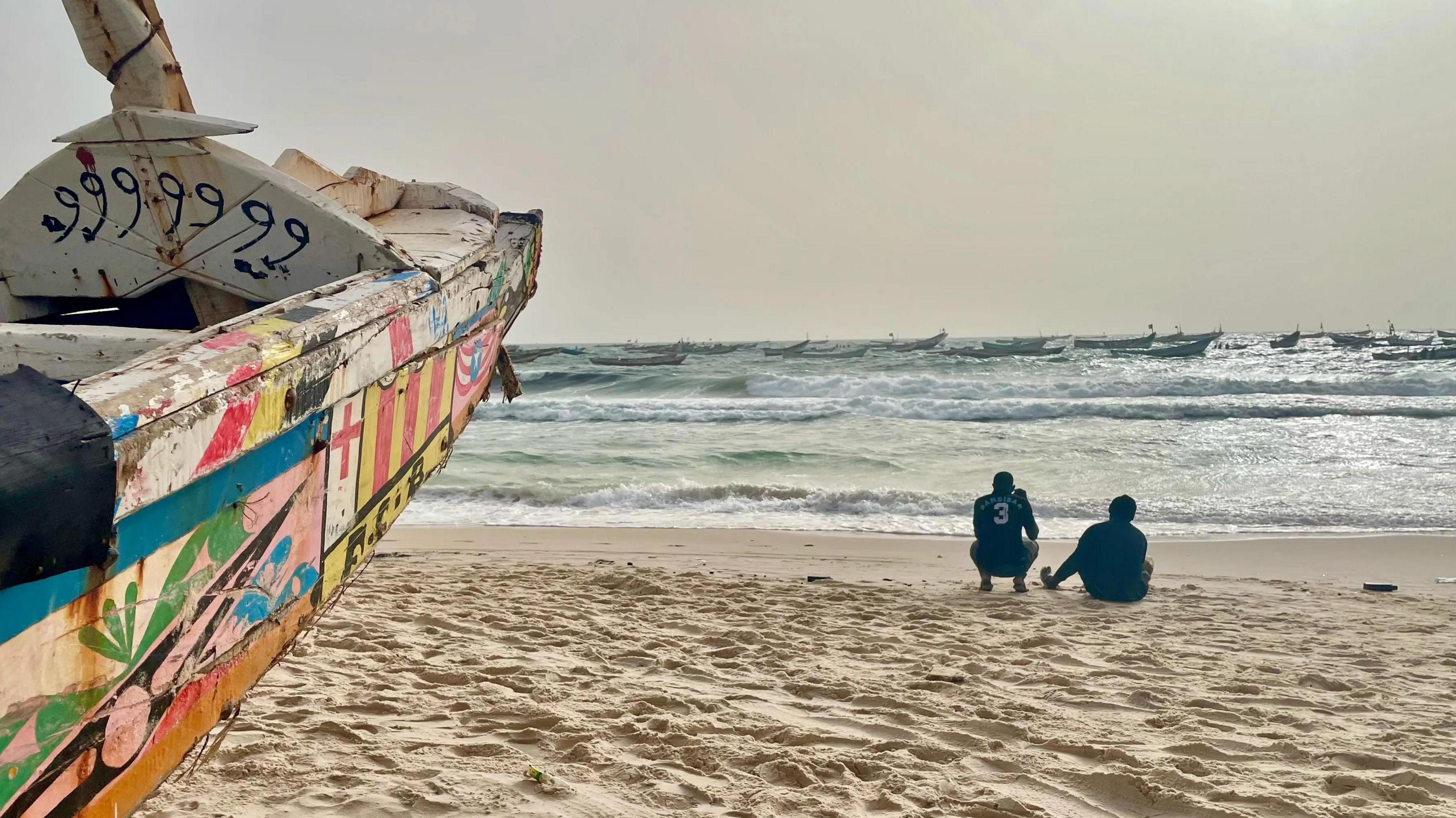 A colourful pirogue to the left on a beach in Mauritania; to the right of the boat two men crouch and look out to sea where other fishing vessels can be seen.