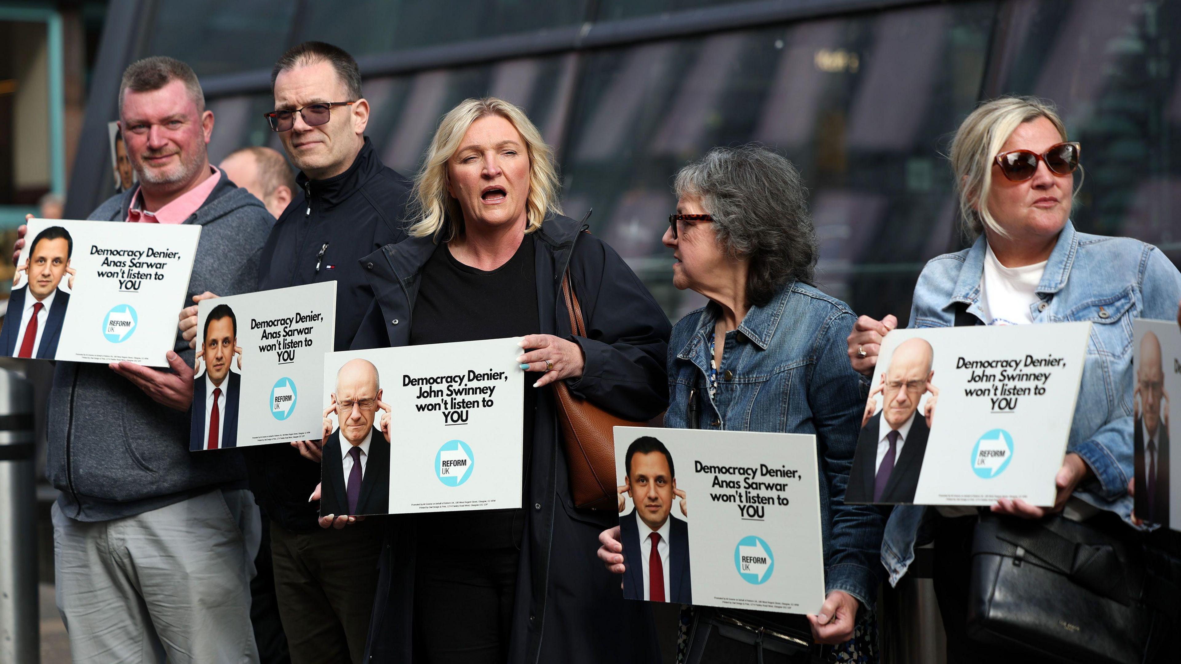 A group of five protestors - two men and three women - holding placards saying "Democracy denier Anas Sarwar won't listen to you" and "Democracy denier John Swinney won't listen to you"