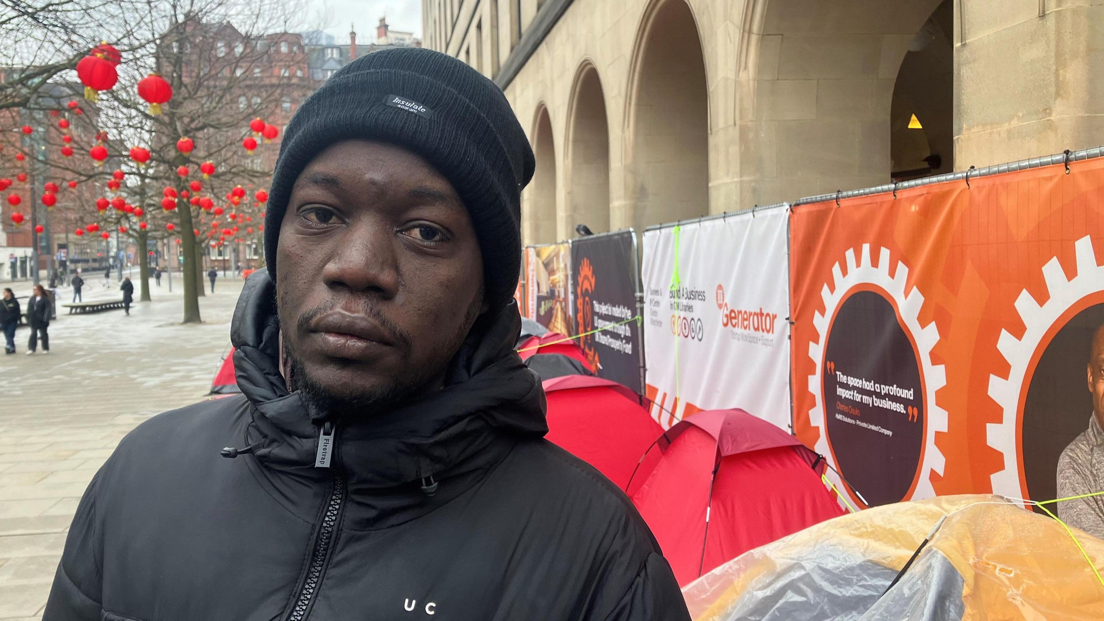 Adam Abdullah wears a black woolly hat and jacket while stood above tents lining the archways of St Peters Square underneath some advertising hoardings. 