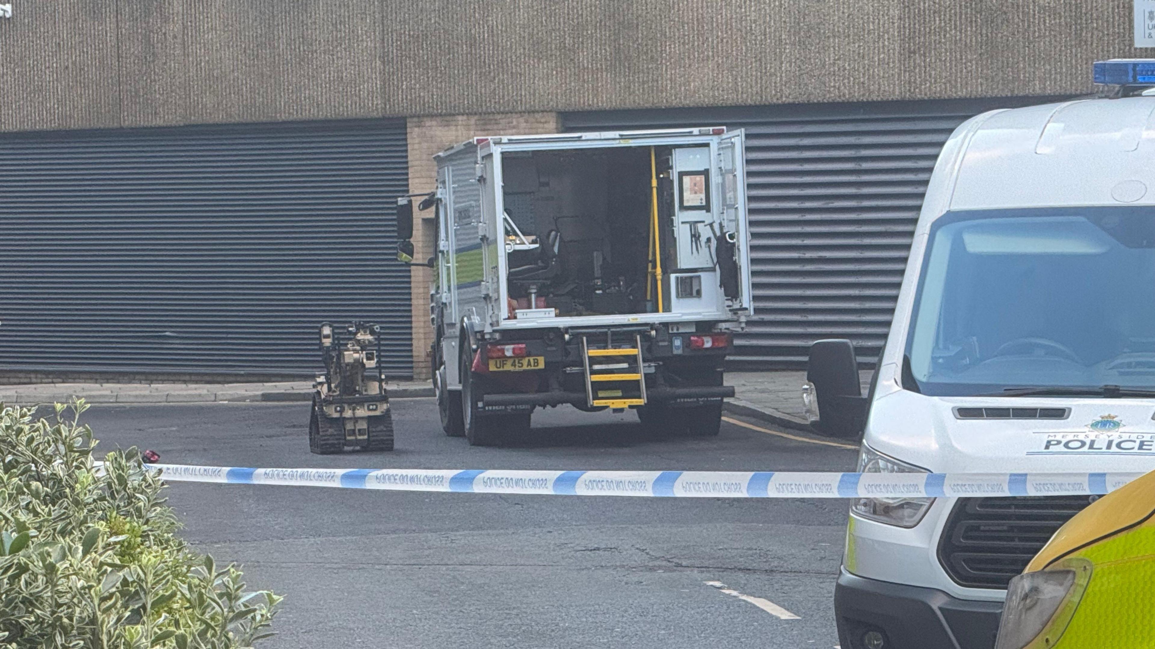 A bomb disposal robot next to a bomb disposal van. The area is blocked off with police tape, the front of a police van can be seen in the foreground.