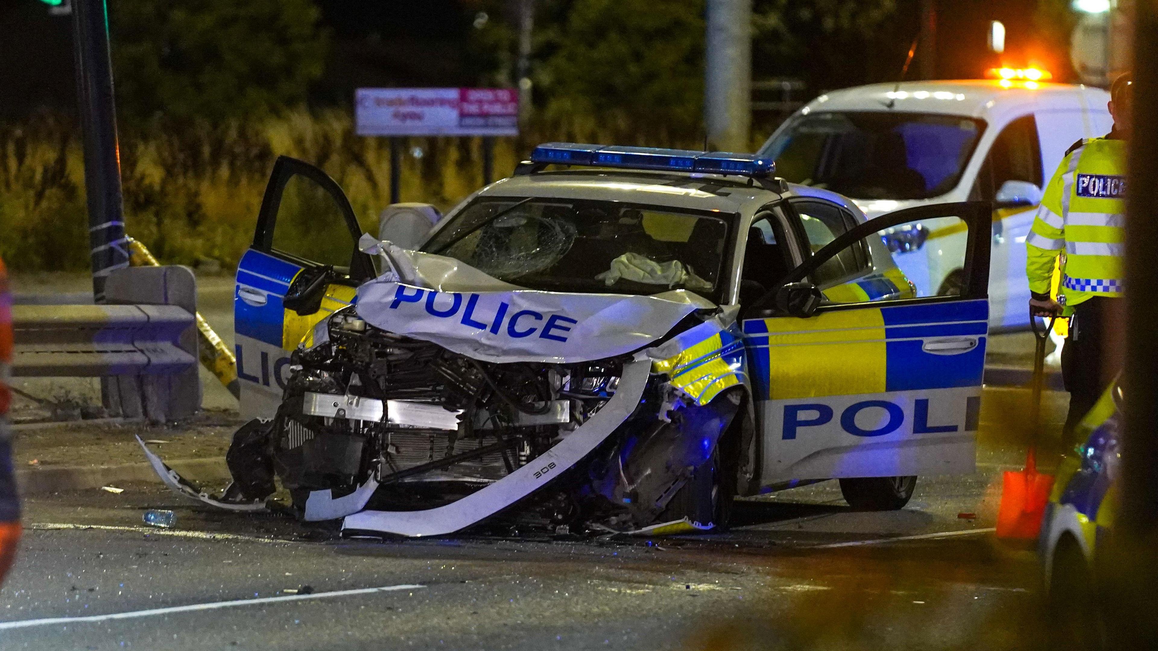 A badly damaged police car with a mangled bonnet and both front doors open. The windscreen is cracked and police officers surround the vehicle.