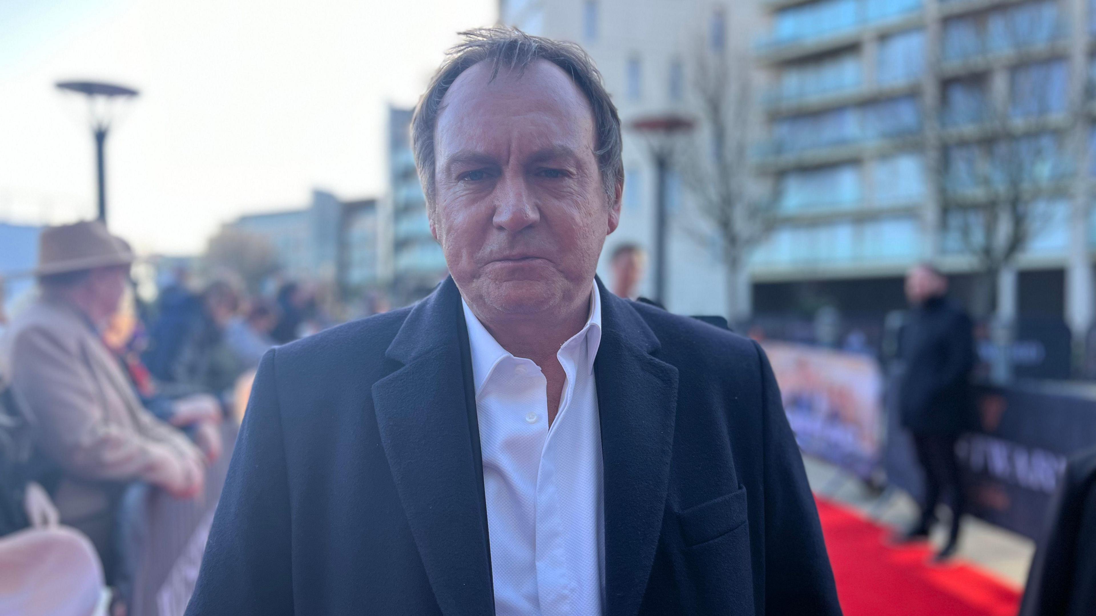 Philip Glenister is looking at the camera while standing on the red carpet. He is wearing a white shirt under a black jacket. He has medium length brown hair. 