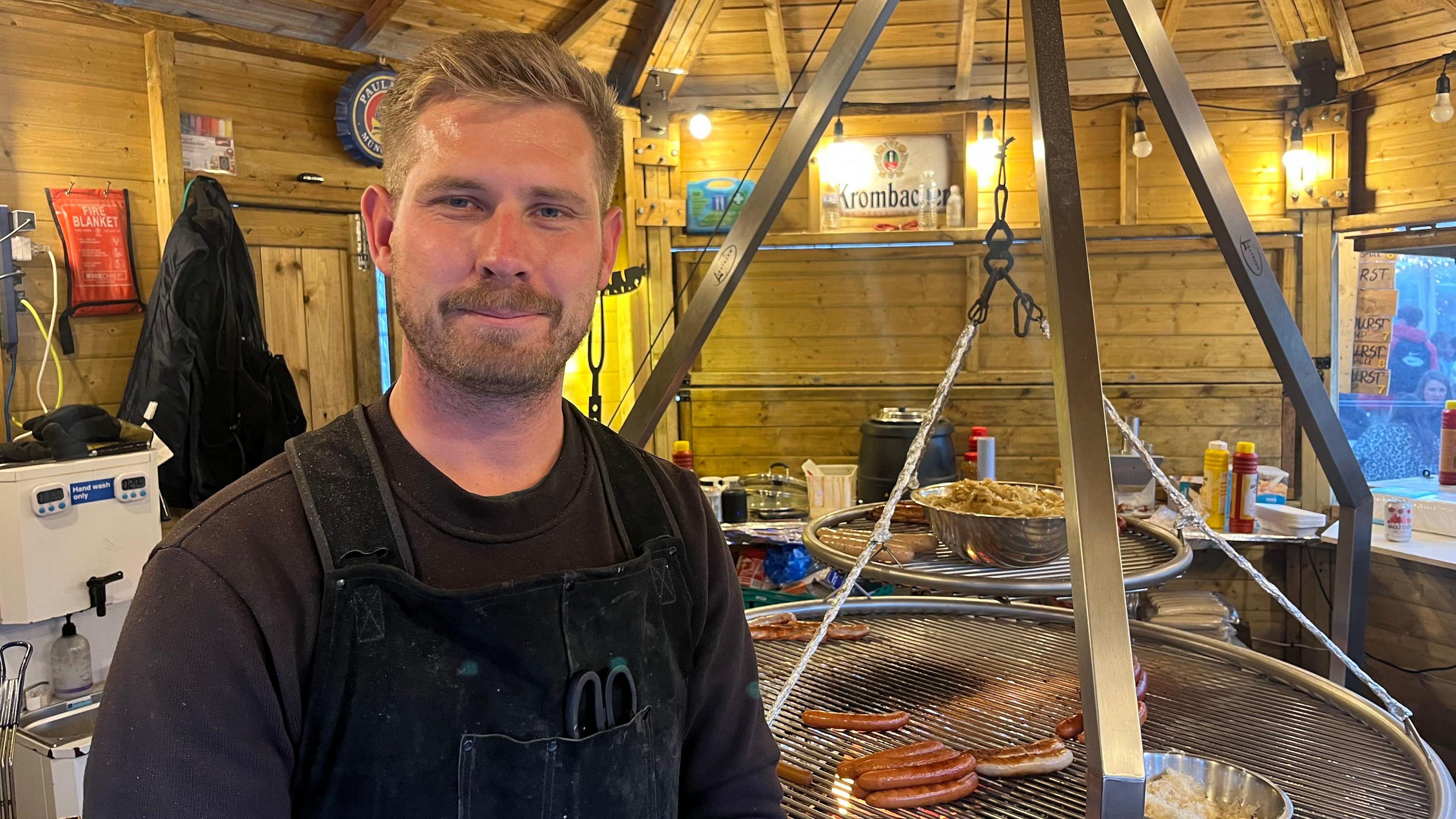 A man standing in a wooden hut with two large circular barbecues with sausages on top. He is wearing a brown top with a black apron over the top.