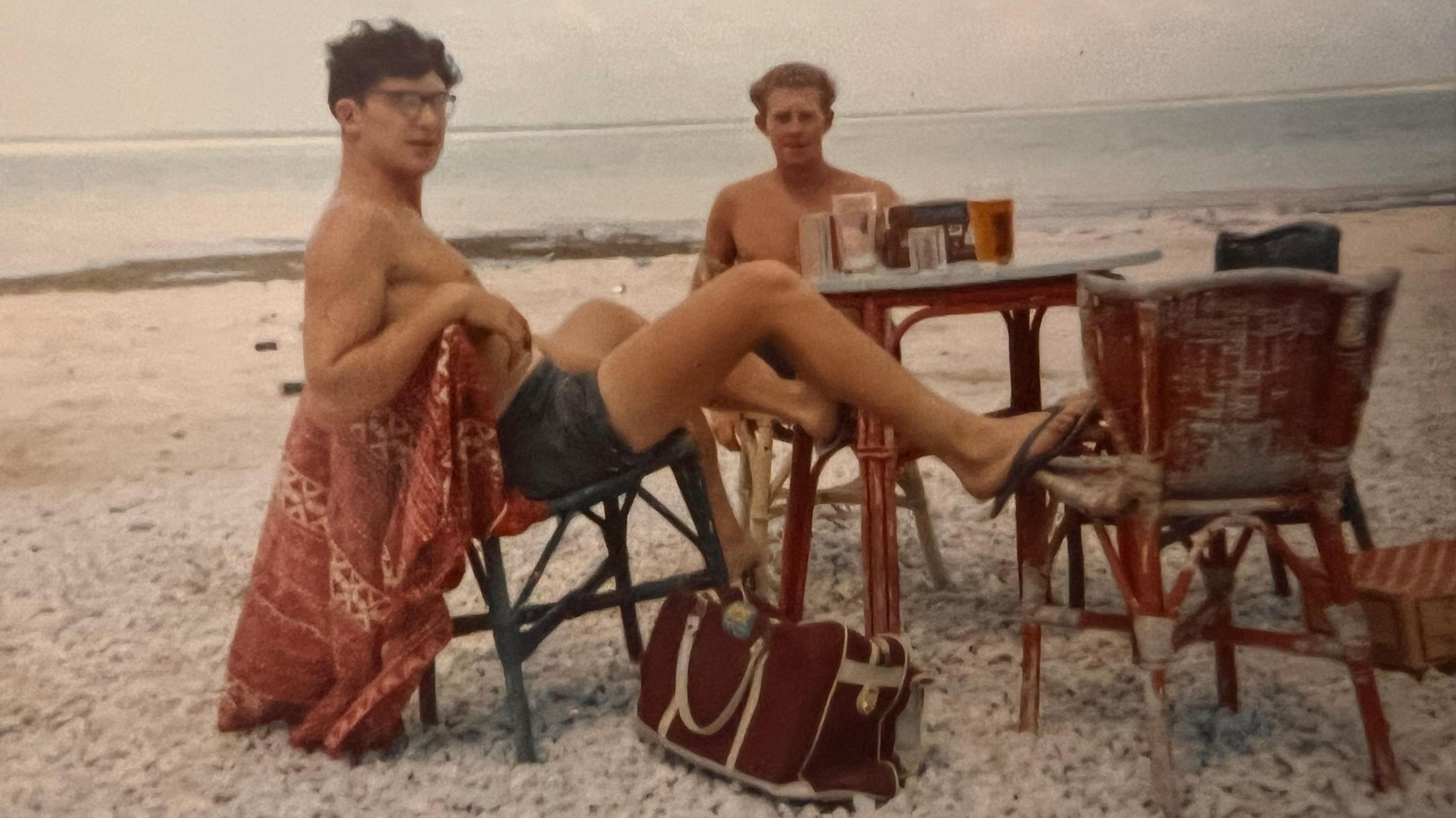 A photograph showing two young men at a table on a beach with the sea in the background and drinks on the table. They are wearing shorts and both looking at the camera.