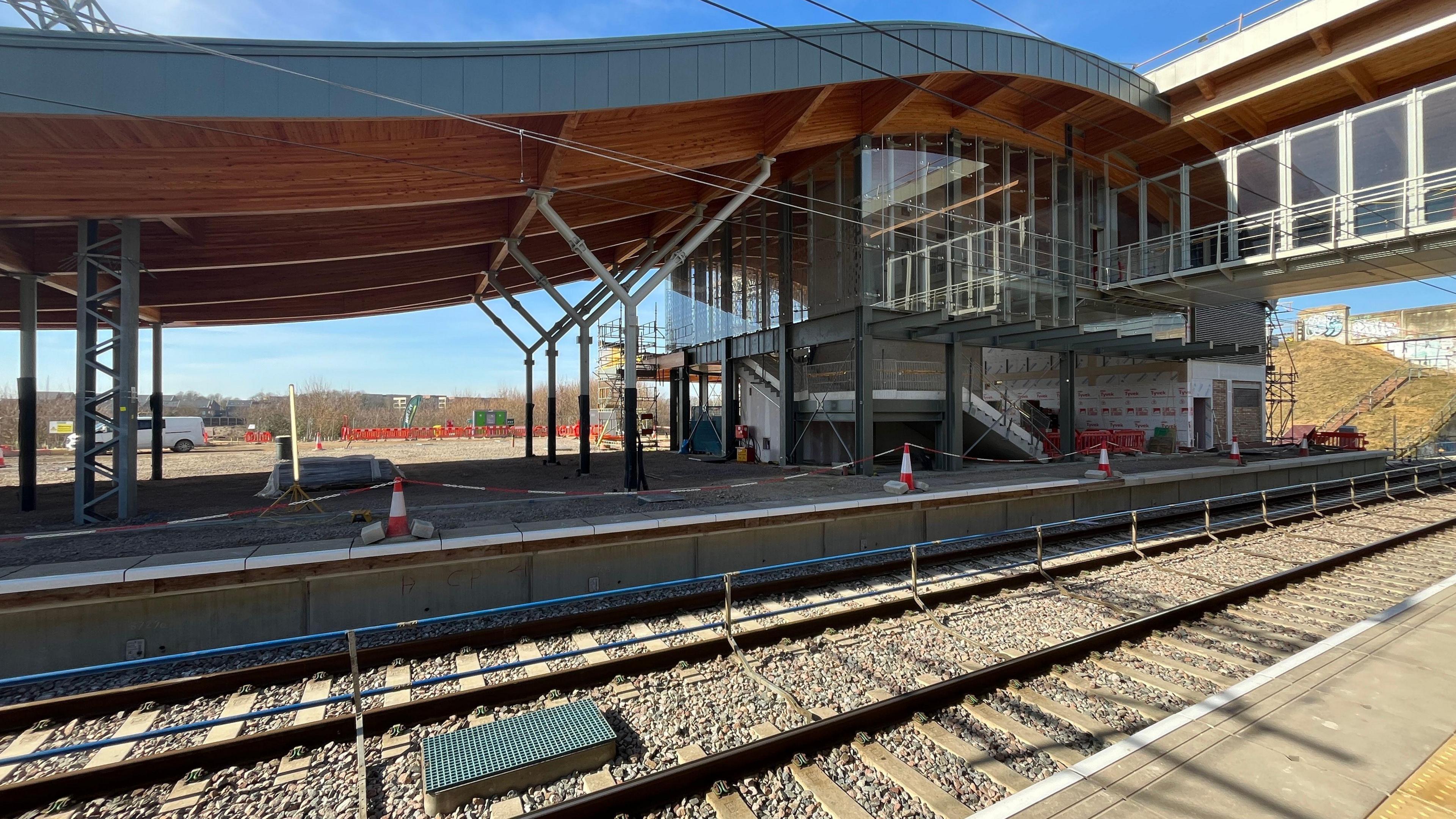 An incomplete Cambridge South railway station. A double train track is in the foreground, with the train station building behind it being constructed. It has a curved roof and there is a walkway going over the top of the tracks. There are also cones and tape blocking off sections on the platform.
