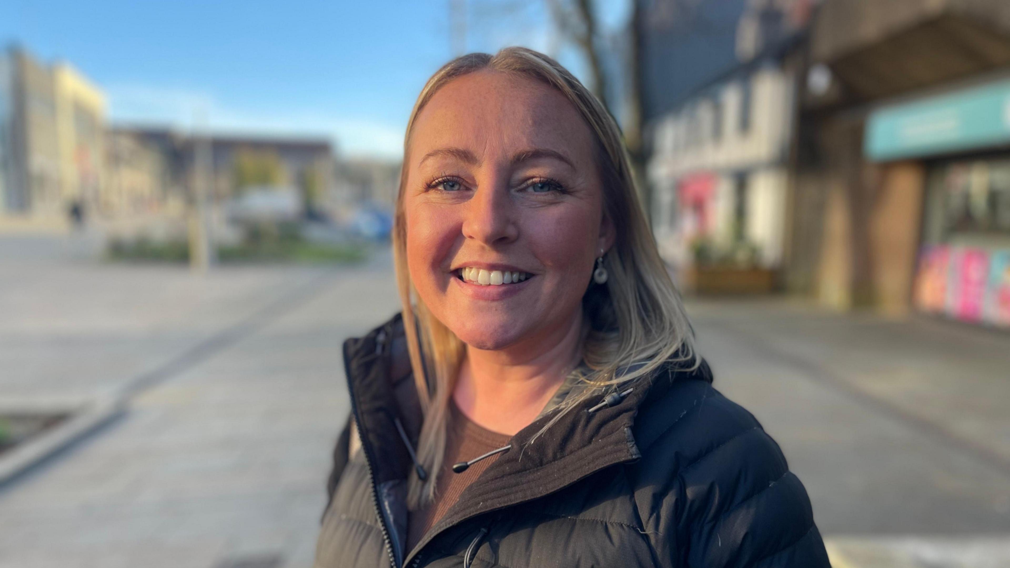 A woman with mid-length blonde hair, wearing a black puffer coat on a shopping street