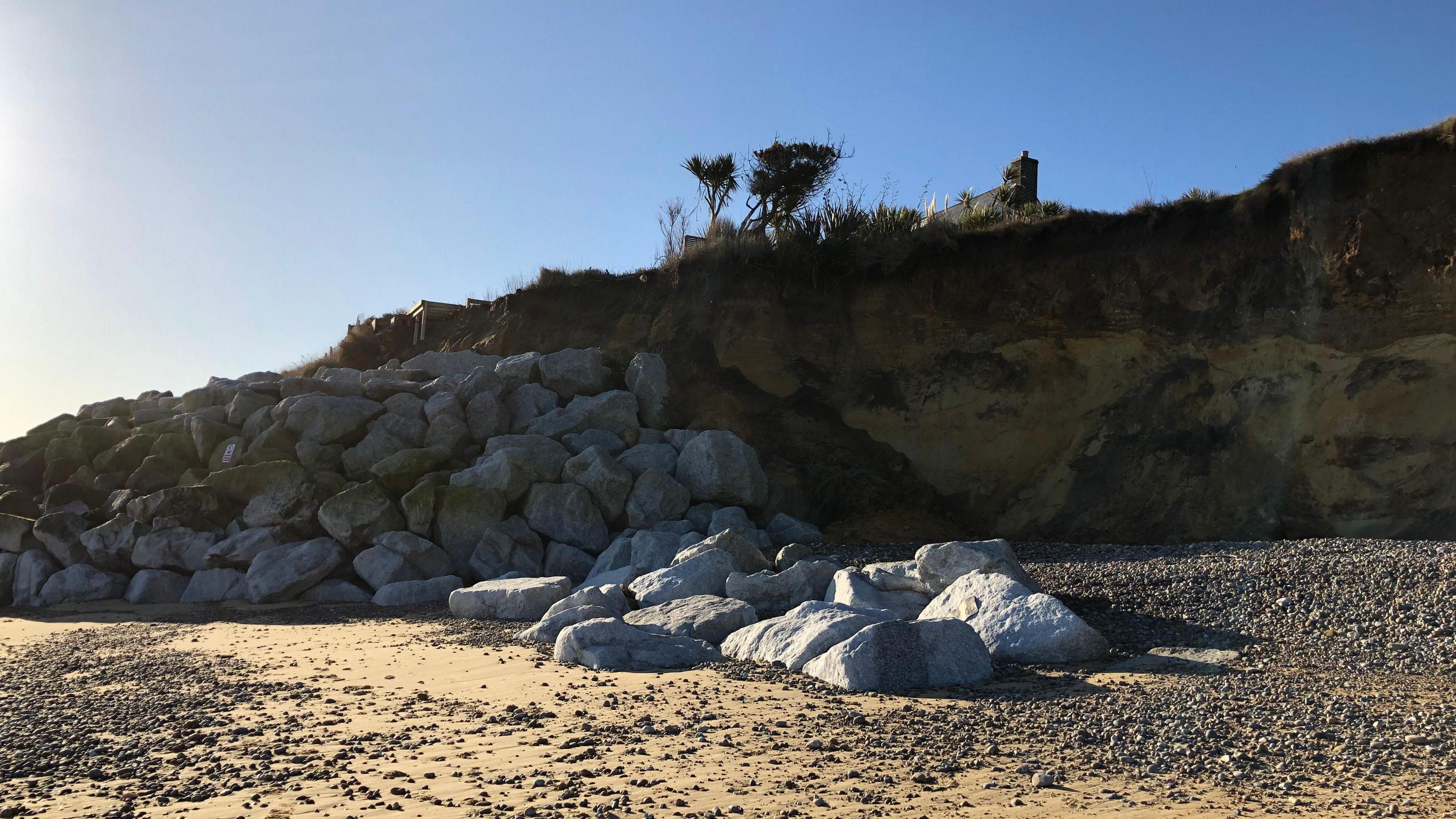 Large boulders are shown against an eroding cliff on a sand and shingle beach with bushes and the chimney of a house seen at the top of the cliff.