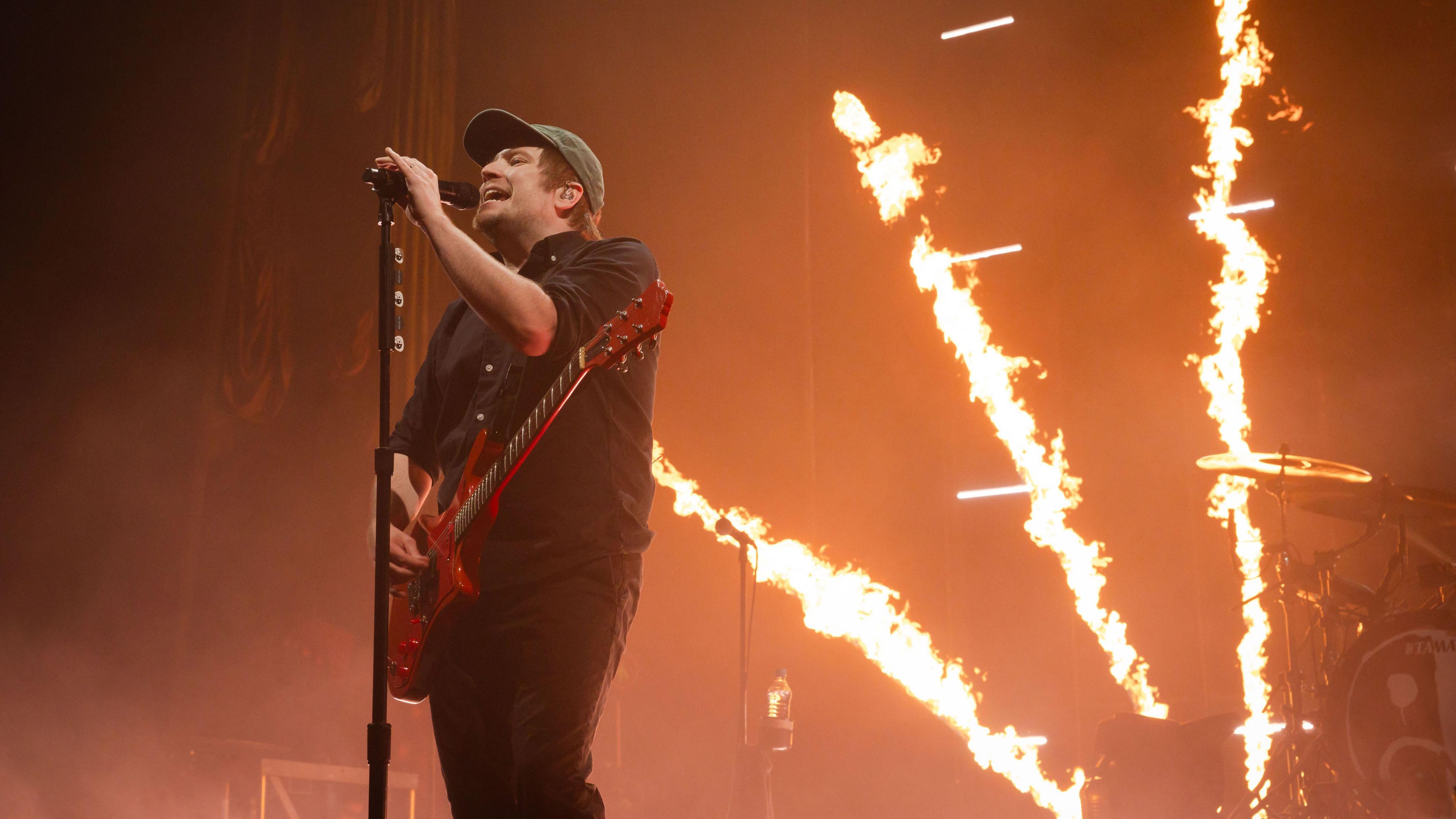 Patrick Stump from the band Fall Out Boy is stood on a stage. He stand behind a microphone stand and wears a black shirt, black jeans and a cap. He also has a red guitar. Behind him, fire shoots up from the stage.