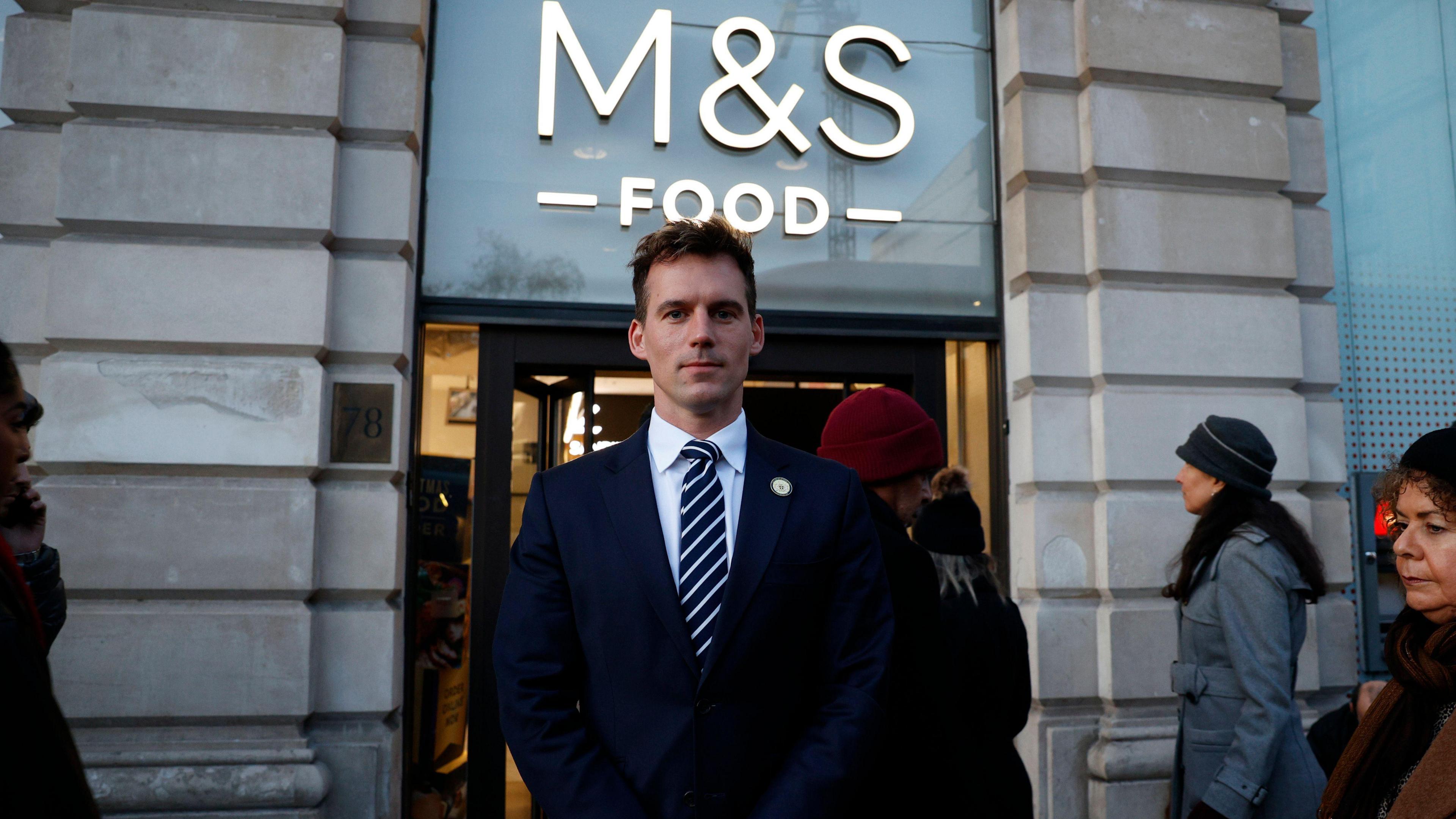 An image of Ed Pitt Ford in a dark blue suit, white shirt and blue and striped tie, standing in front of the entrance to the M&S Food store in Green Park. People mill around him in the background