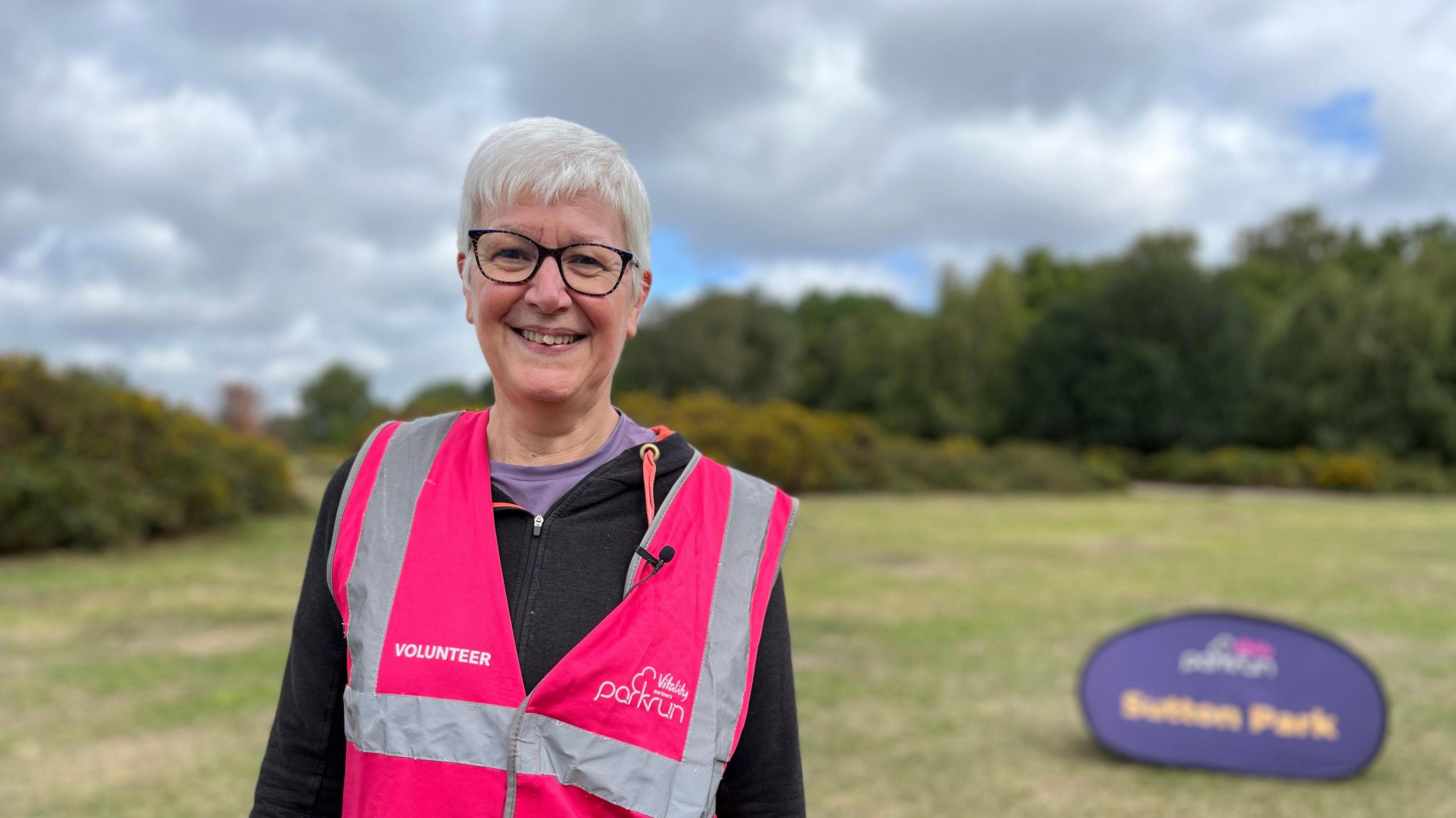 Parkrun volunteer Sue Garton stands in Sutton Park with a pink parkrun bib on top of a black fleece. She has short white hair and wears glasses and is smiling at camera.
