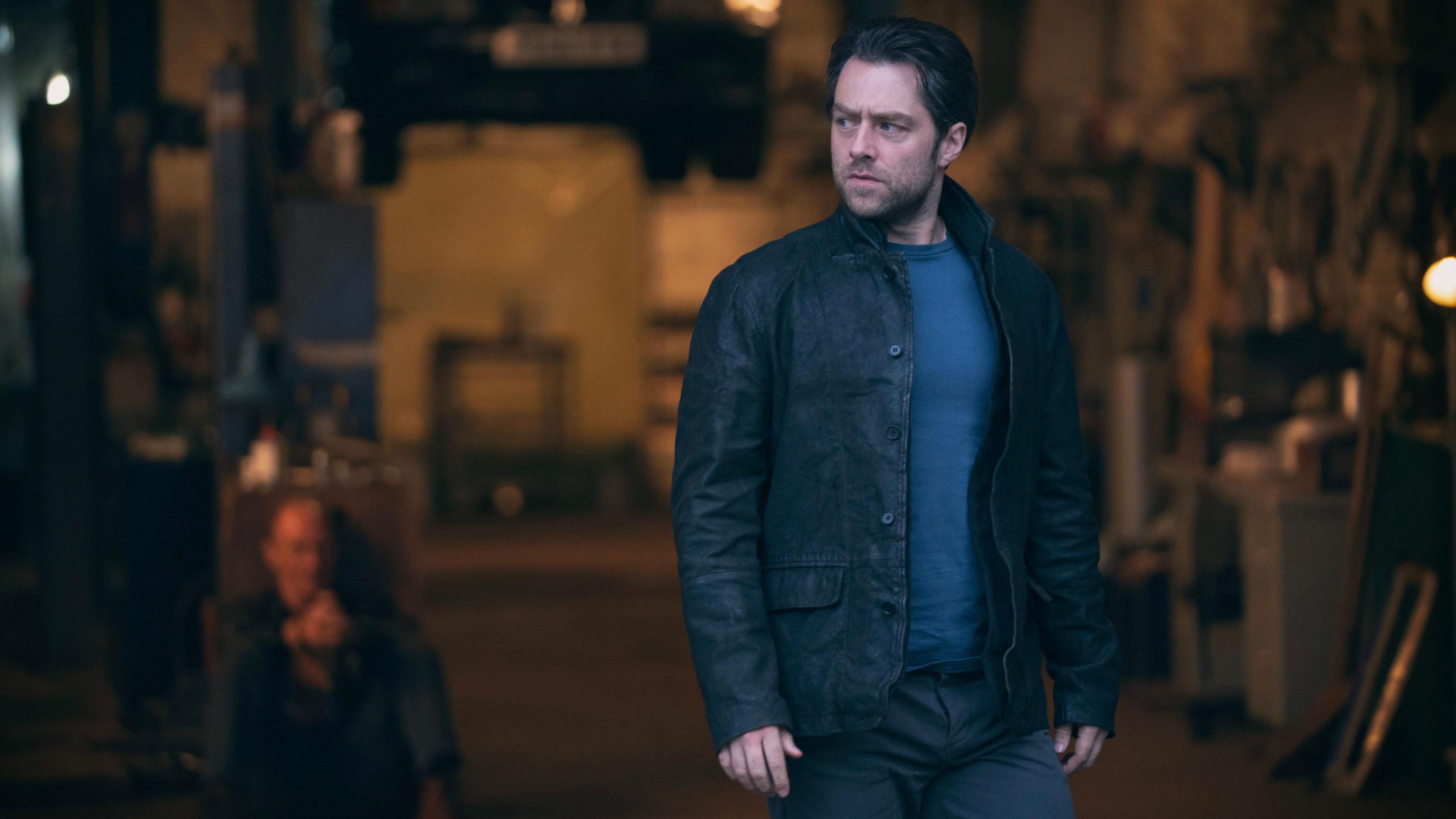 Richard Rankin, a dark-haired, brooding man in leather jacket and T-shirt looks seriously off camera. Behind him, in what appears to be a garage, a bald man is sitting on the floor, his hands apparently bound together.