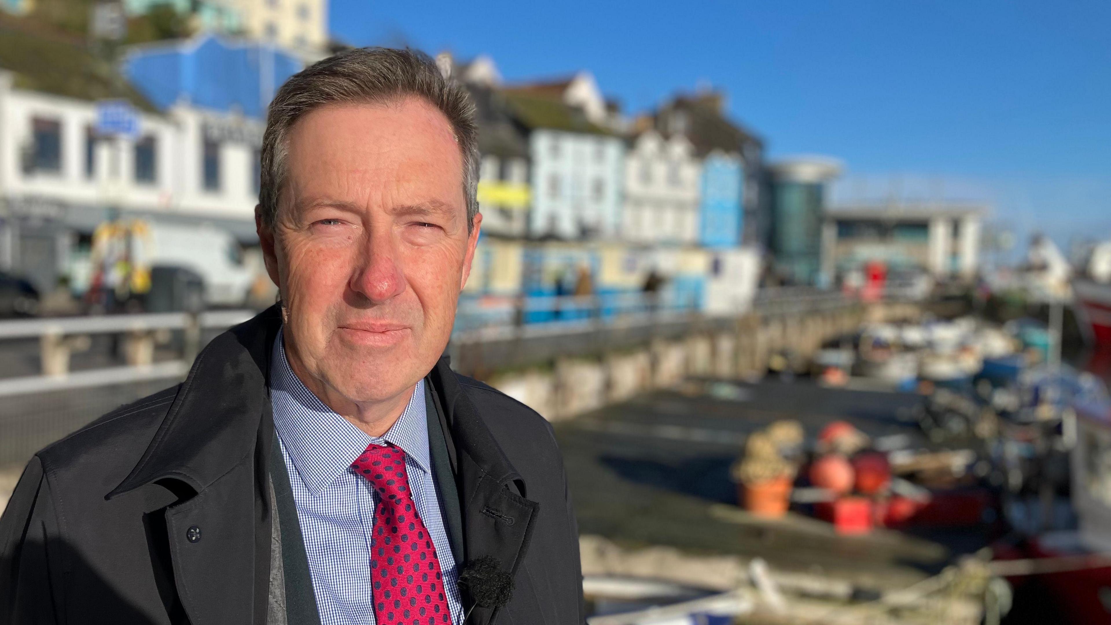 A man stands by a harbour, with colourful shop fronts and houses in the background.  You see boats and trawlers in the water. 