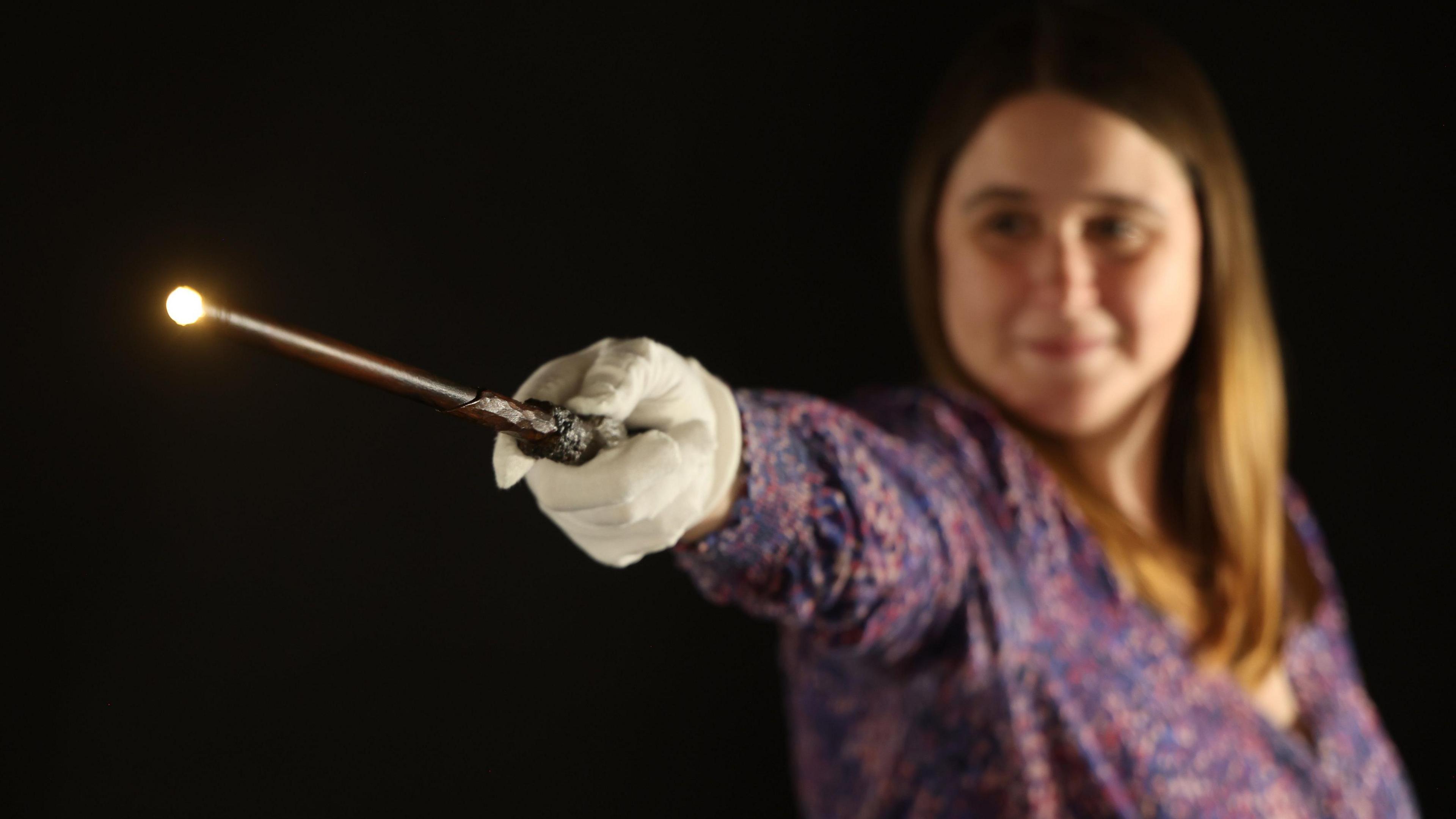 A woman wearing a purple floral top with long light hair is pointing the wand which is lit at the top with her arm fully outstretched