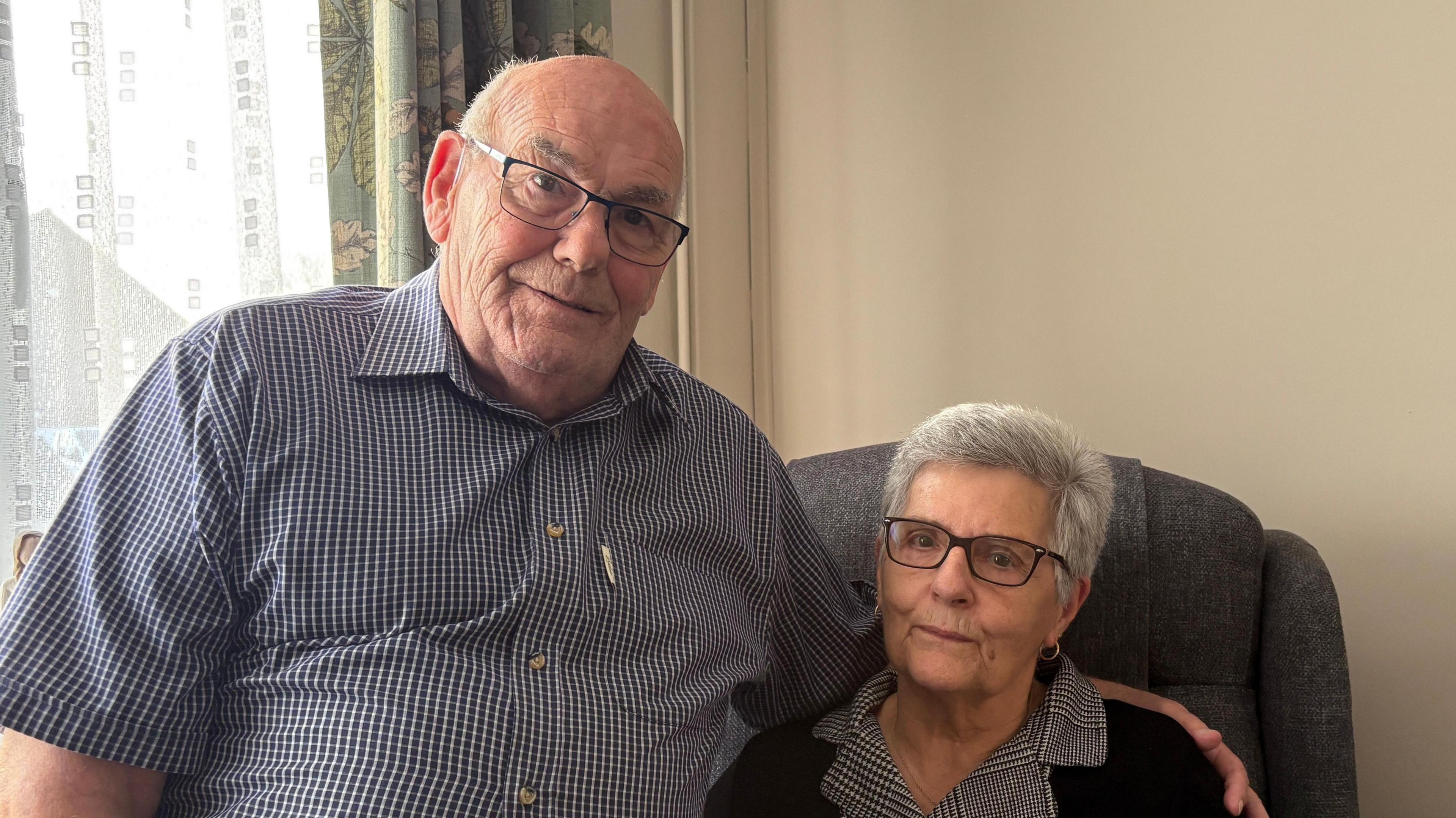 Terrence and Lesley Bridges, looking down the camera and sitting down inside. Lesley, on the right, is seated in a grey armchair. Terrence, on the left, is sitting next to her, on the arm of the chair. Behind Lesley is a blank cream-coloured wall, while behind Terrence is a window covered in net curtains, which are closed, and light blue fabric curtains, which are open. Terrence is wearing a short-sleeved blue checked shirt, which is open at the collar, and black-rimmed rectangular glasses. He is bald on the top of his head, with some short grey hair at the sides. Lesley is wearing a black and white collared top under a black cardigan, as well as black-rimmed rectangular glasses. She has greying hair.