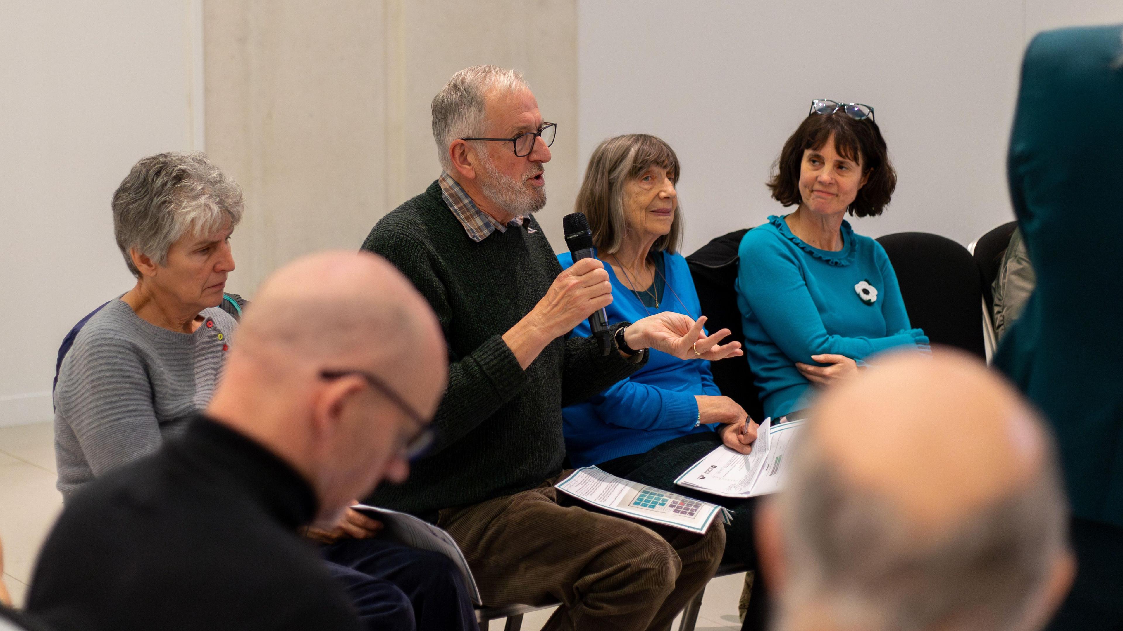 Three women and one man are sat on chairs facing to the right. The man is wearing glasses and speaking into a microphone while gesturing with his other hand. He has some papers on his knee. Two men are sat facing him in the foreground.