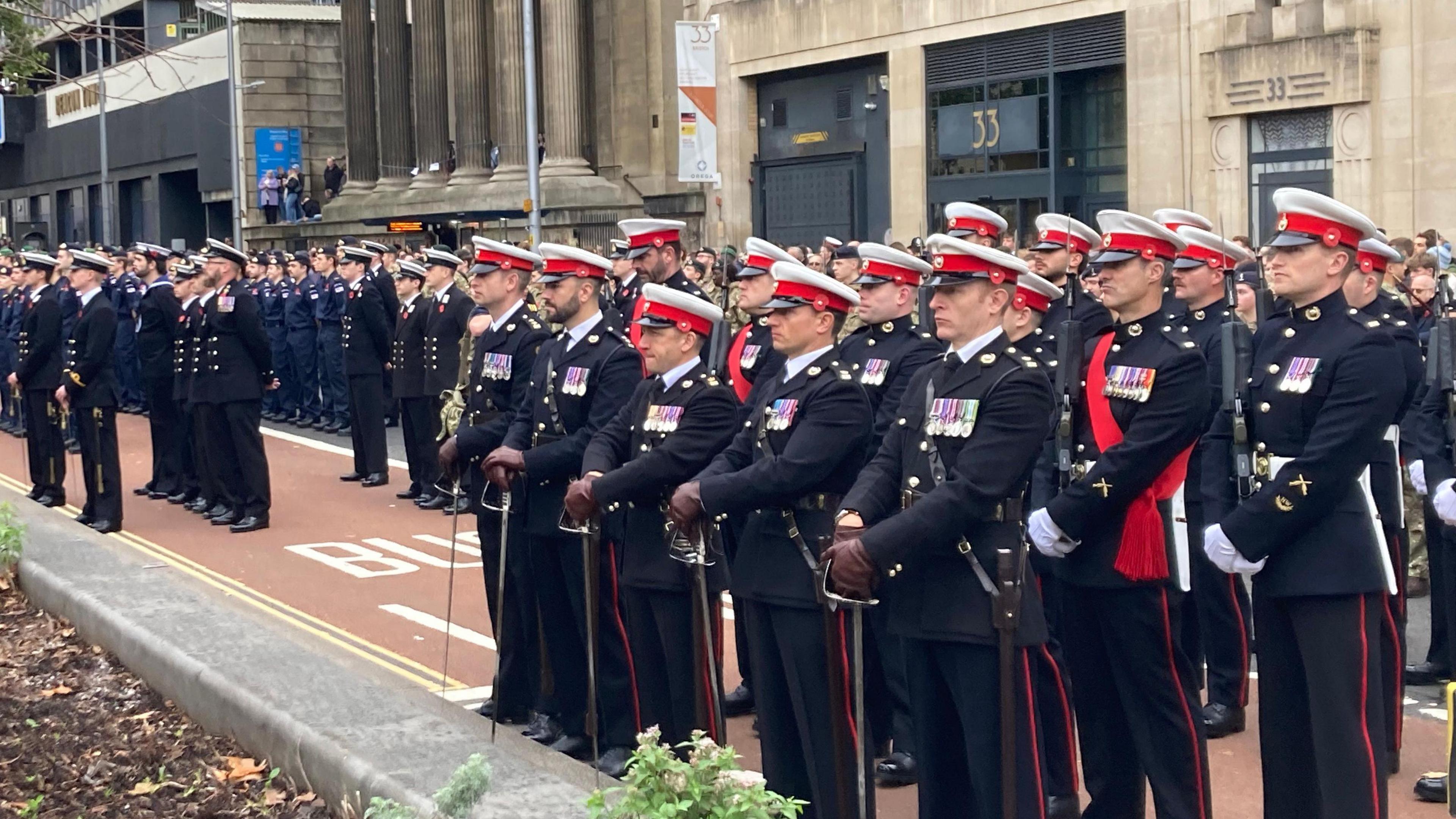 A group of men in military uniform. They are all wearing black suits and hats that are red and white. Many of them have medals on their jackets too. They are all standing in rows and looking forward.
