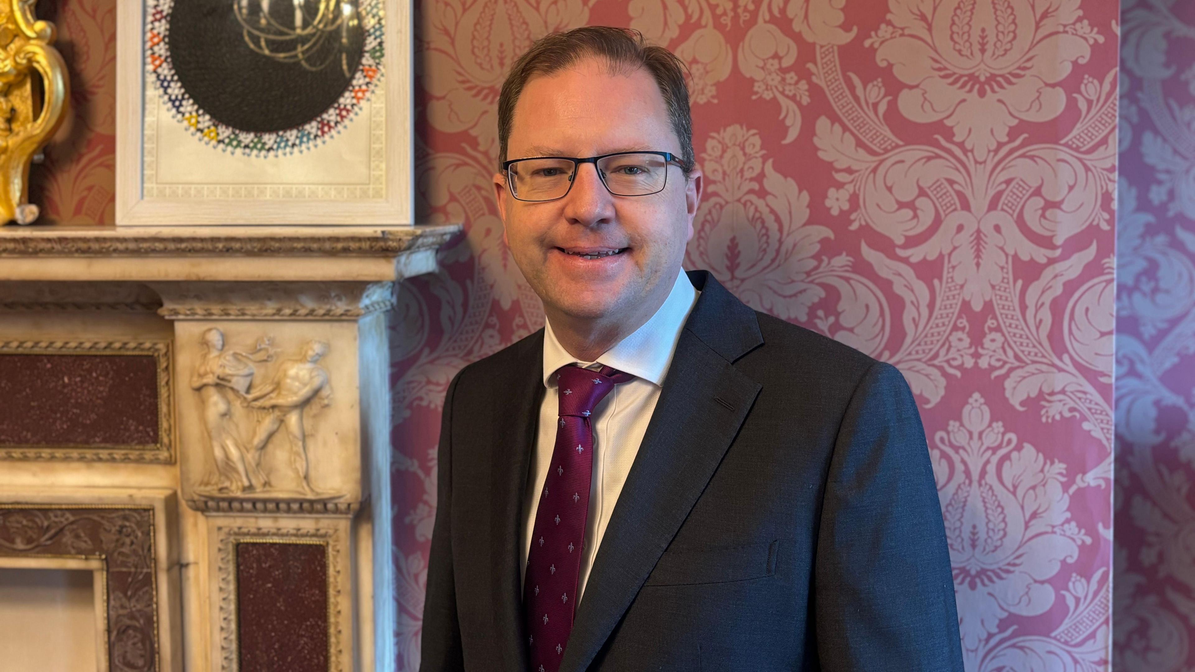 Irish government minister who is a man wearing a suit smiles at the camera from his office