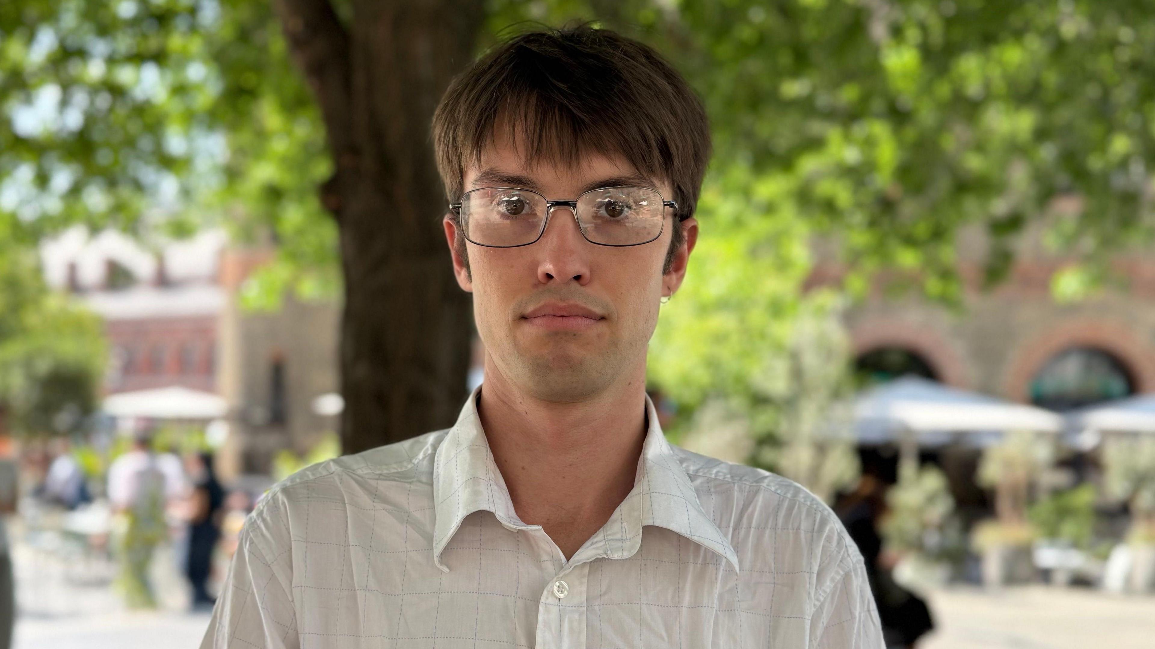 Paddy Friend in a white shirt with a thin check pattern. He has brown hair and glasses.