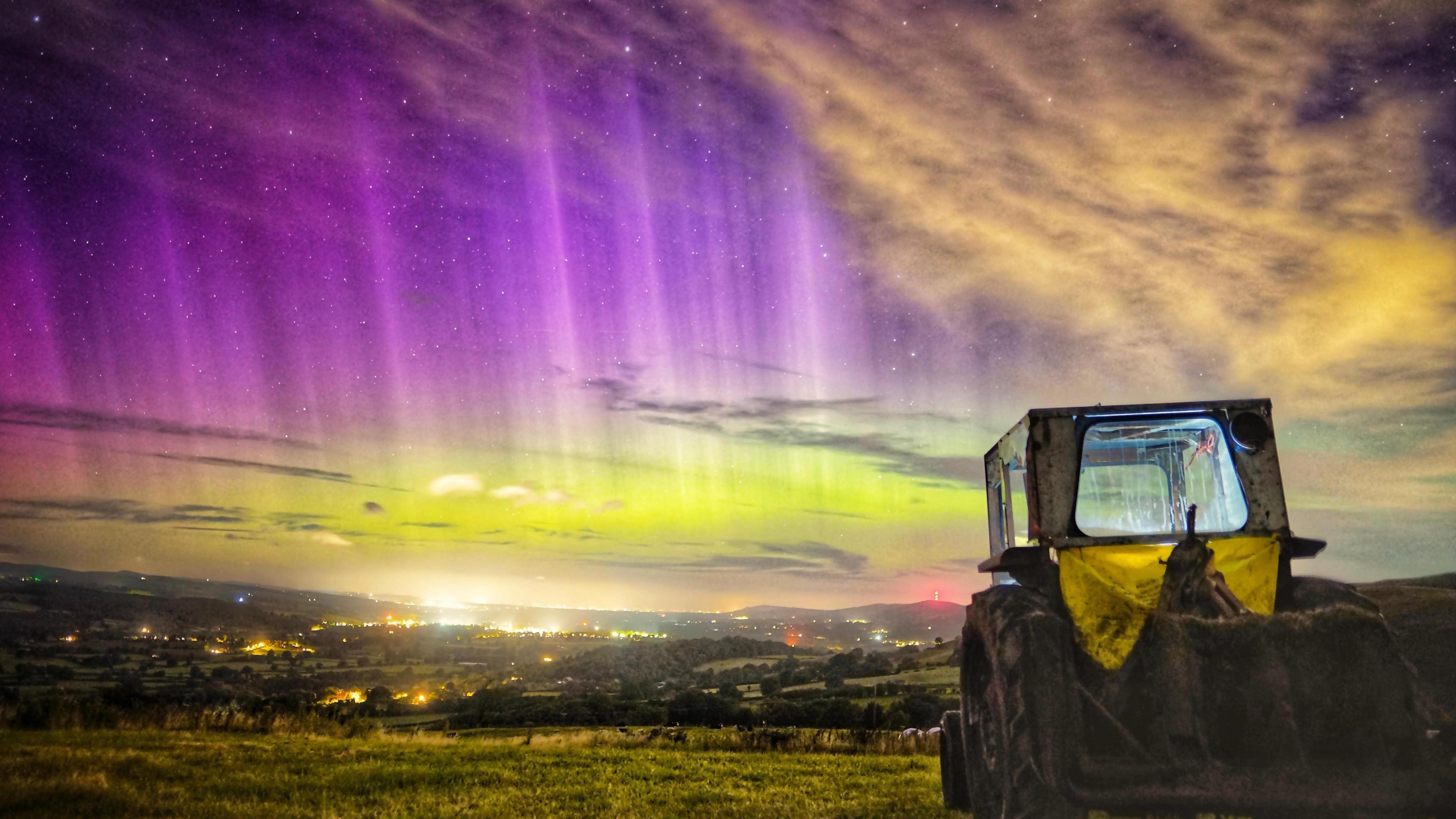 a view across a valley with an old vintage tractor in the foreground.  Behind it the night sky is filled with green and purple colours of the northern lights with some shafts of white light