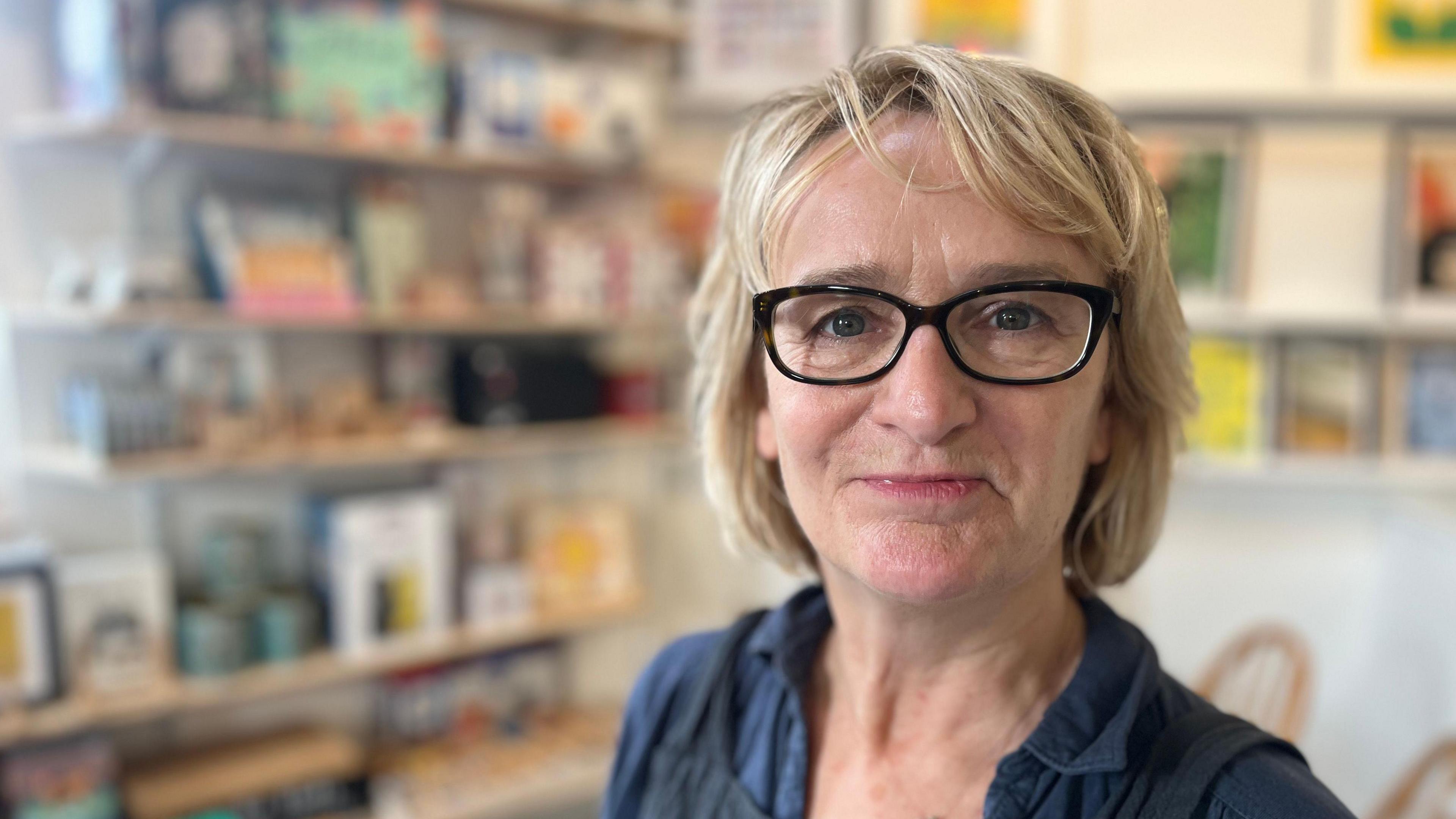 Justine Hyde, owner of Hyde and Seek, wearing black-framed glasses and a navy shirt against a blurred background of gifts.