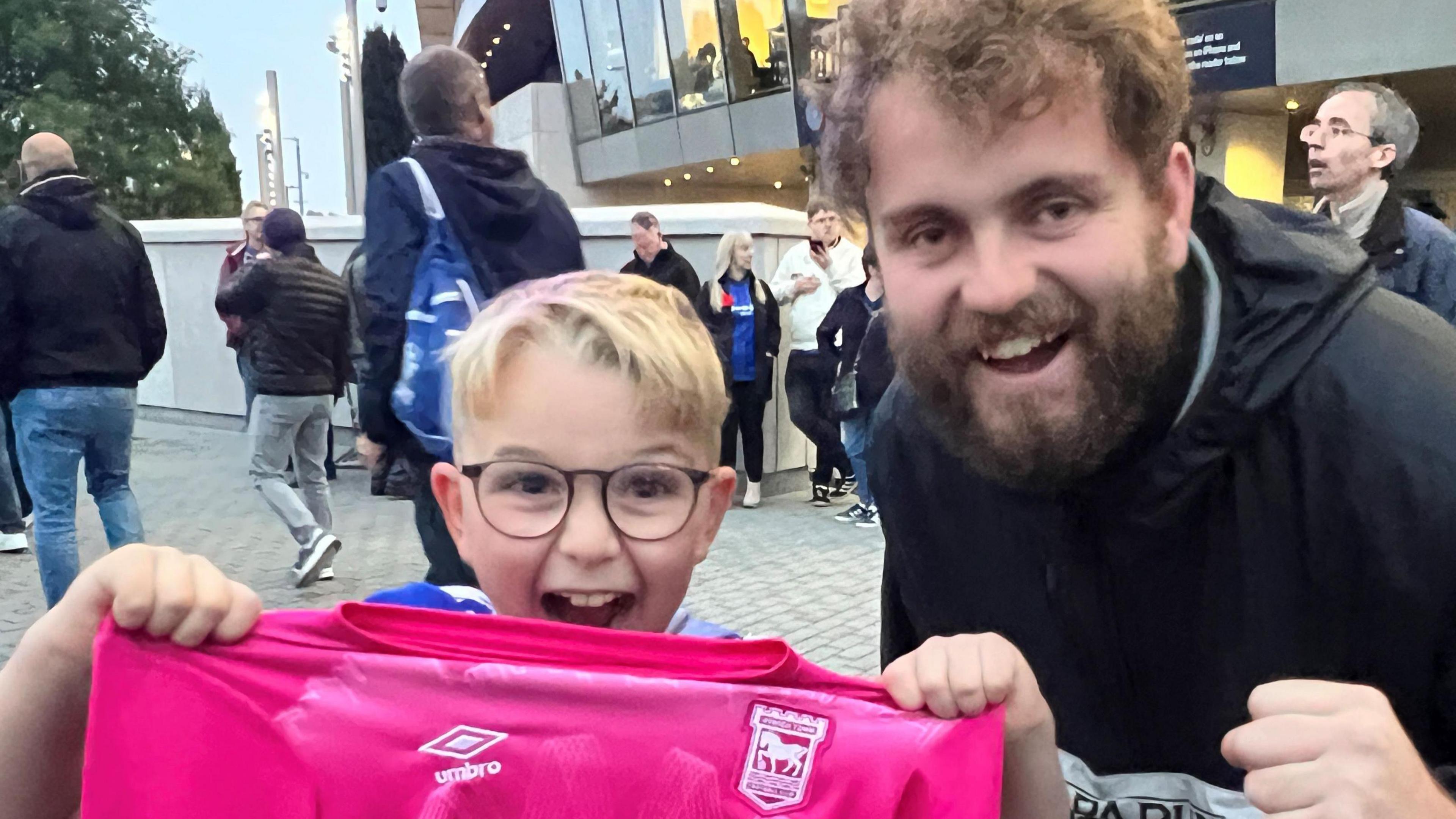 A dad and son are centre. The boy is around 10 and had short blonde hair.  He's wearing glasses and is holding up a pink ITFC third kit shirt.  His dad has dark brown hair and a beard and moustache.  He's wearing a black hooded jacket. In the background other Town fans are mingling outside the Tottenham Hotspur Stadium.