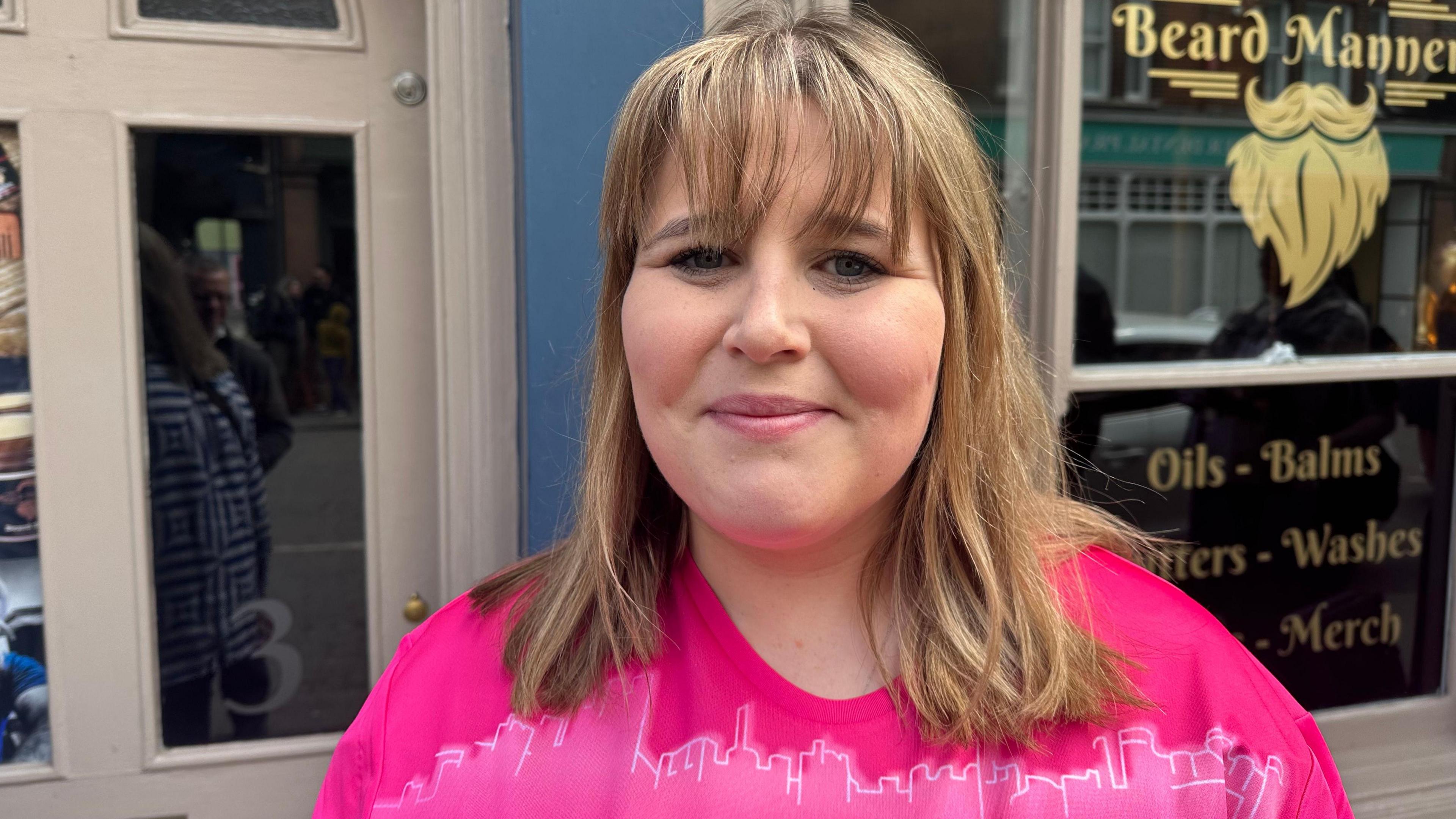 Woman with blond hair wears a pink T-shirt and stands with her back to a pub