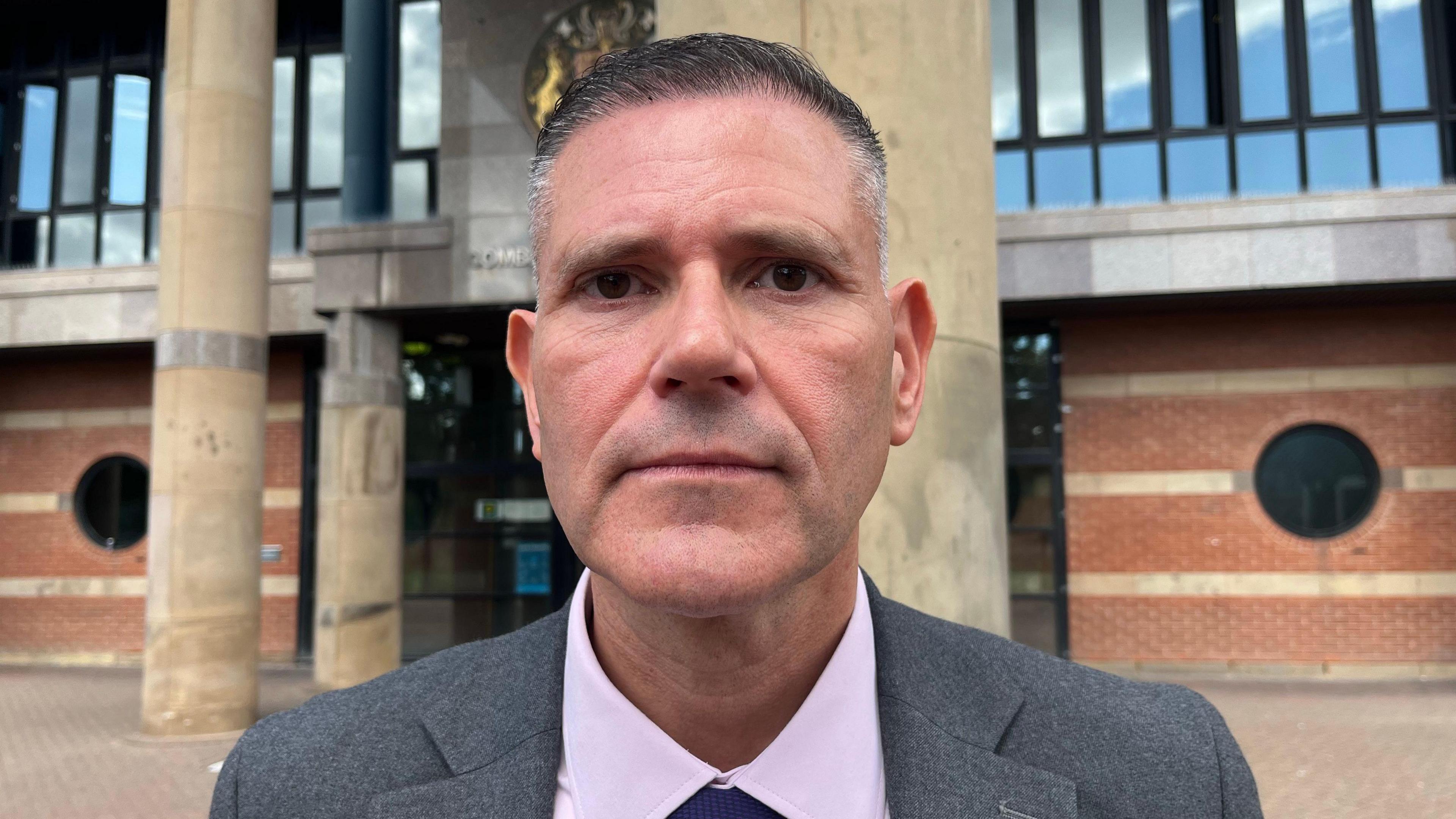 Steve Chatterton outside Teesside Crown Court. He is clean shaven with neat short dark greying hair, and is wearing a grey suit with a pink shirt and blue tie. His expression is sombre and serious.