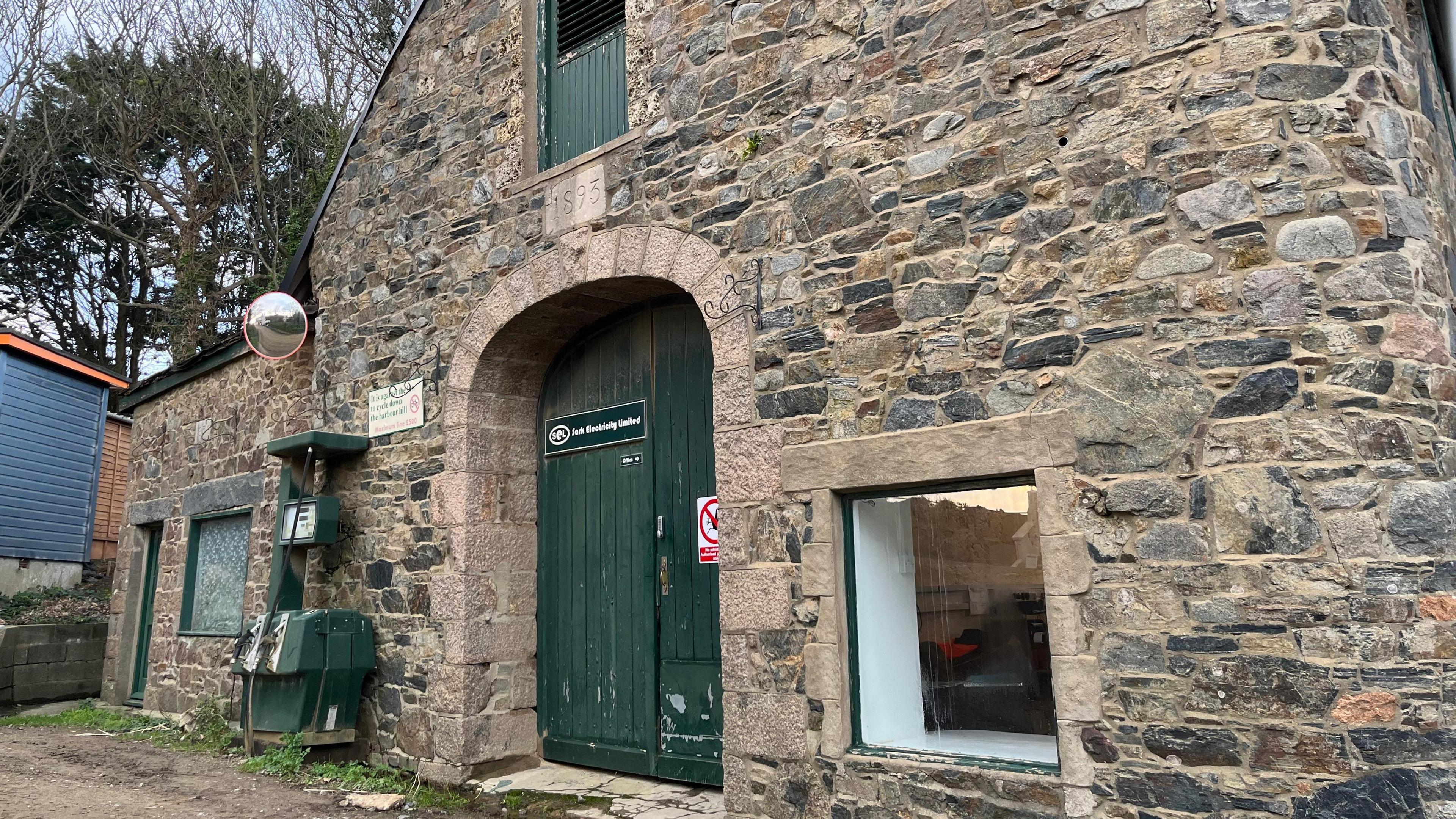 A brick building with a green door and a window. A sign says Sark Electricity. A petrol pump in the same green is also attached to the brick wall. 