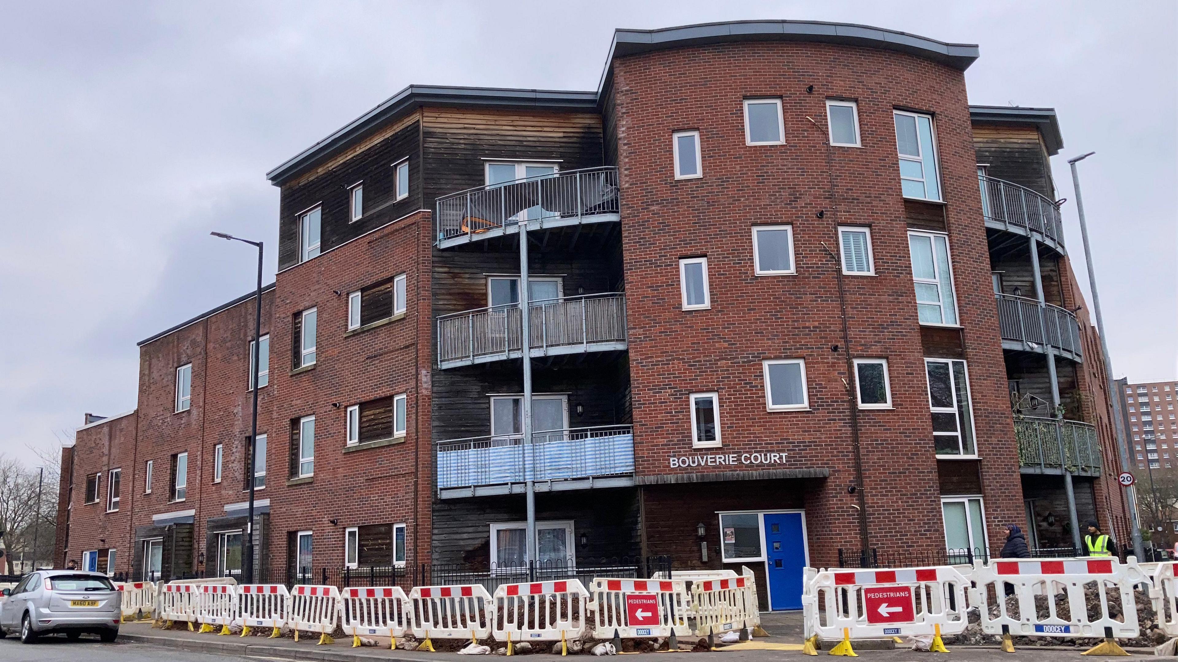 An exterior shot of Bouverie Court, it is a red brick building with a mix of terraced housing to the left and a three-story block of flats with large balconies on the right hand side of the building. There are roadworks going on on the pavement outside