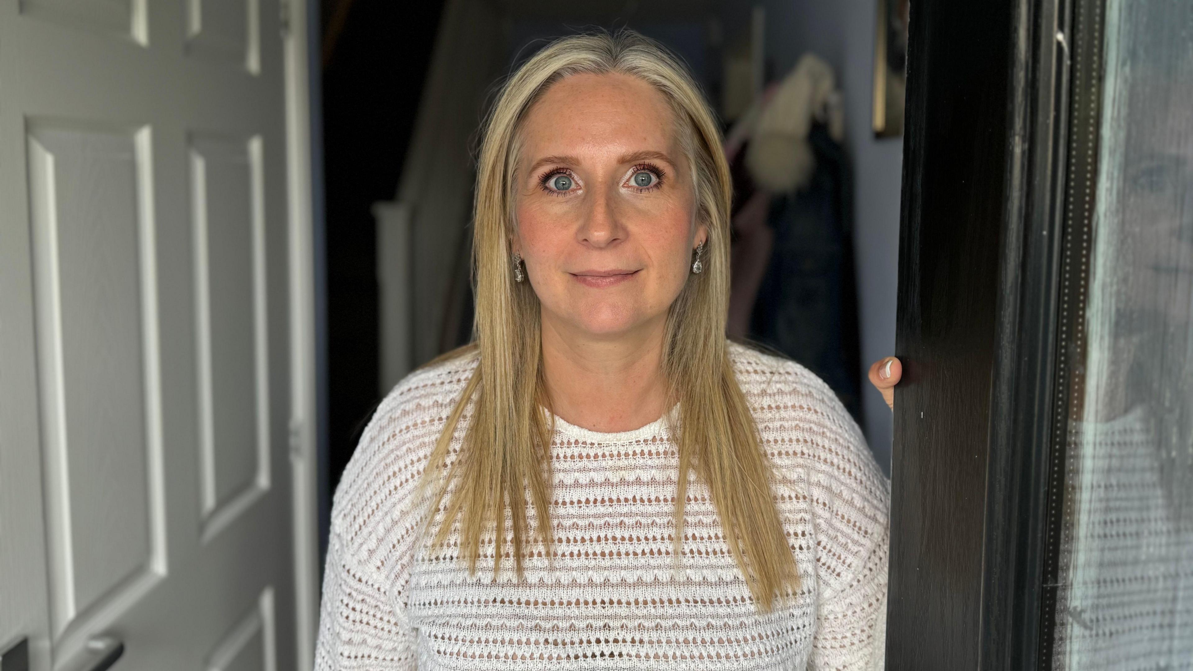 Natalie Braine with long blonde hair and wearing a white top looking directly into the camera and standing in her doorway. 