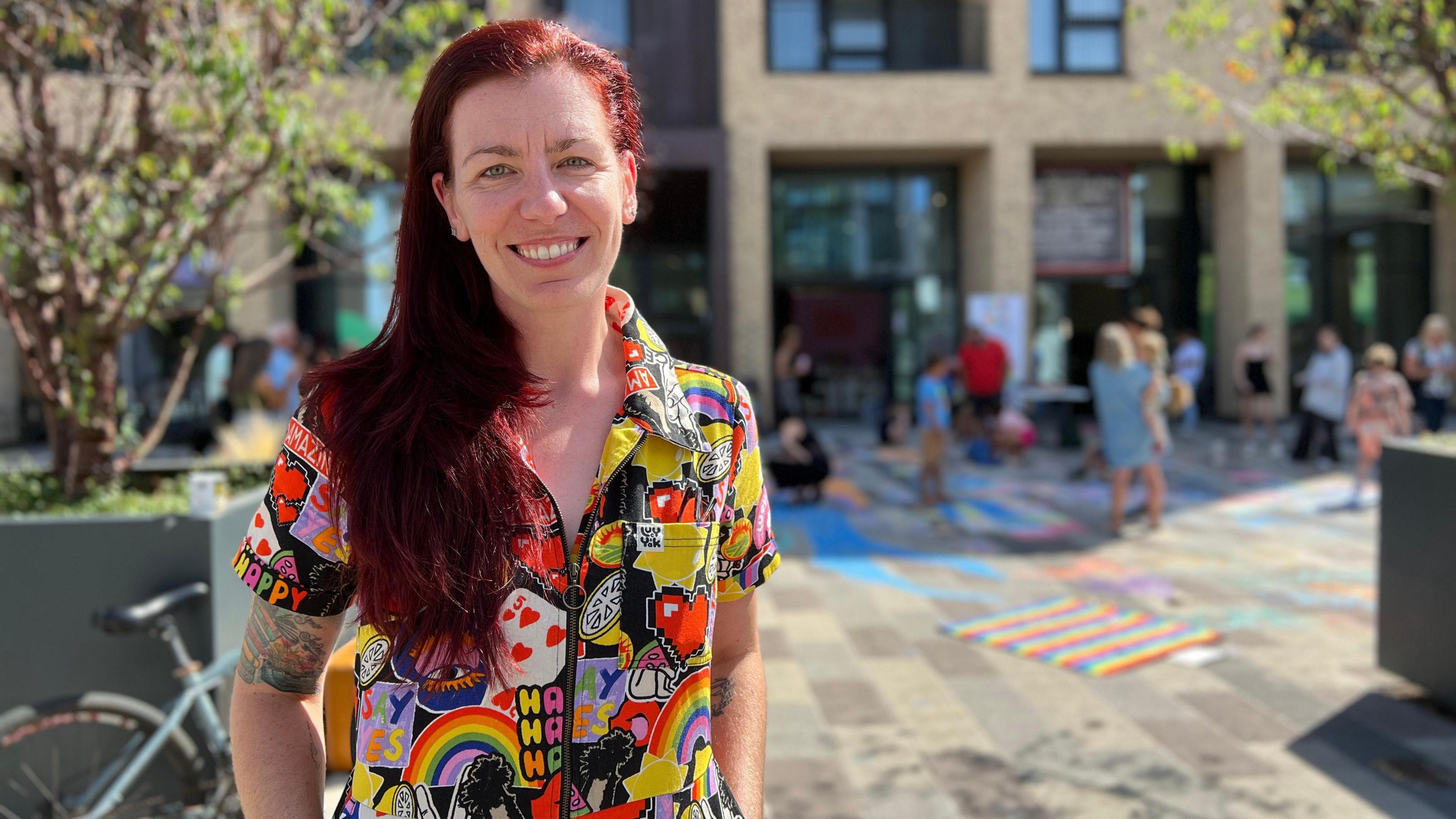 Kaye stands outdoors in front of a modern building, wearing a colorful shirt with rainbows, hearts, and the word 'HAPPY' repeated across it. The paved area around them is decorated with chalk drawings, and people are walking or sitting nearby. Trees and a bicycle are visible to the left