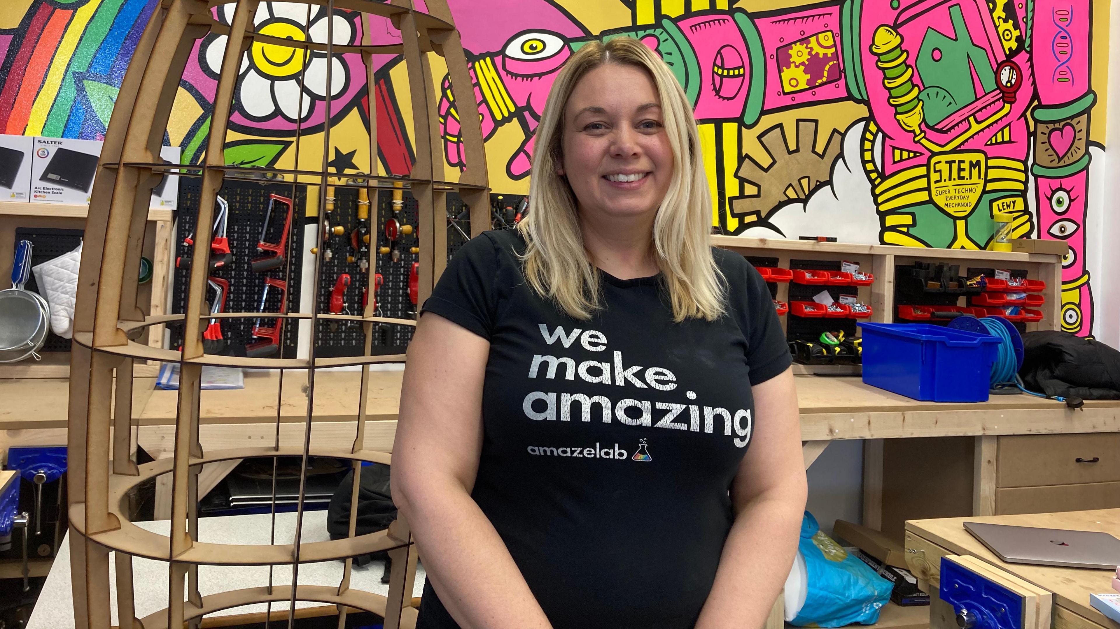 Mrs Briggs stands in front of a colourful wall which features shelves filled with tools. She wears a black t-shirt with the words "we make amazing" in white letters on the front and the business logo below.