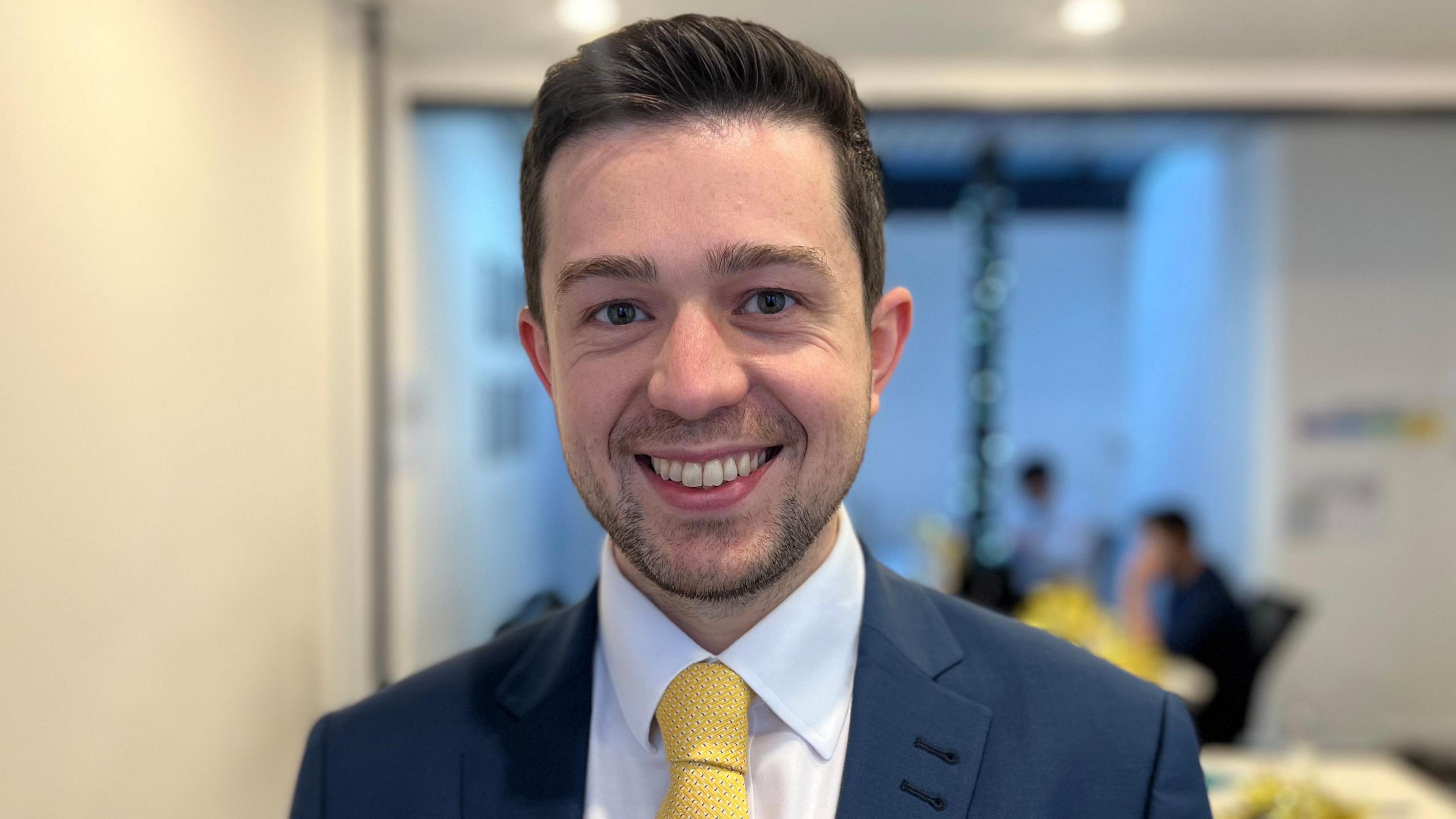 Head and shoulders of George Heritage wearing a white collar shirt, yellow tie and blue jacket.