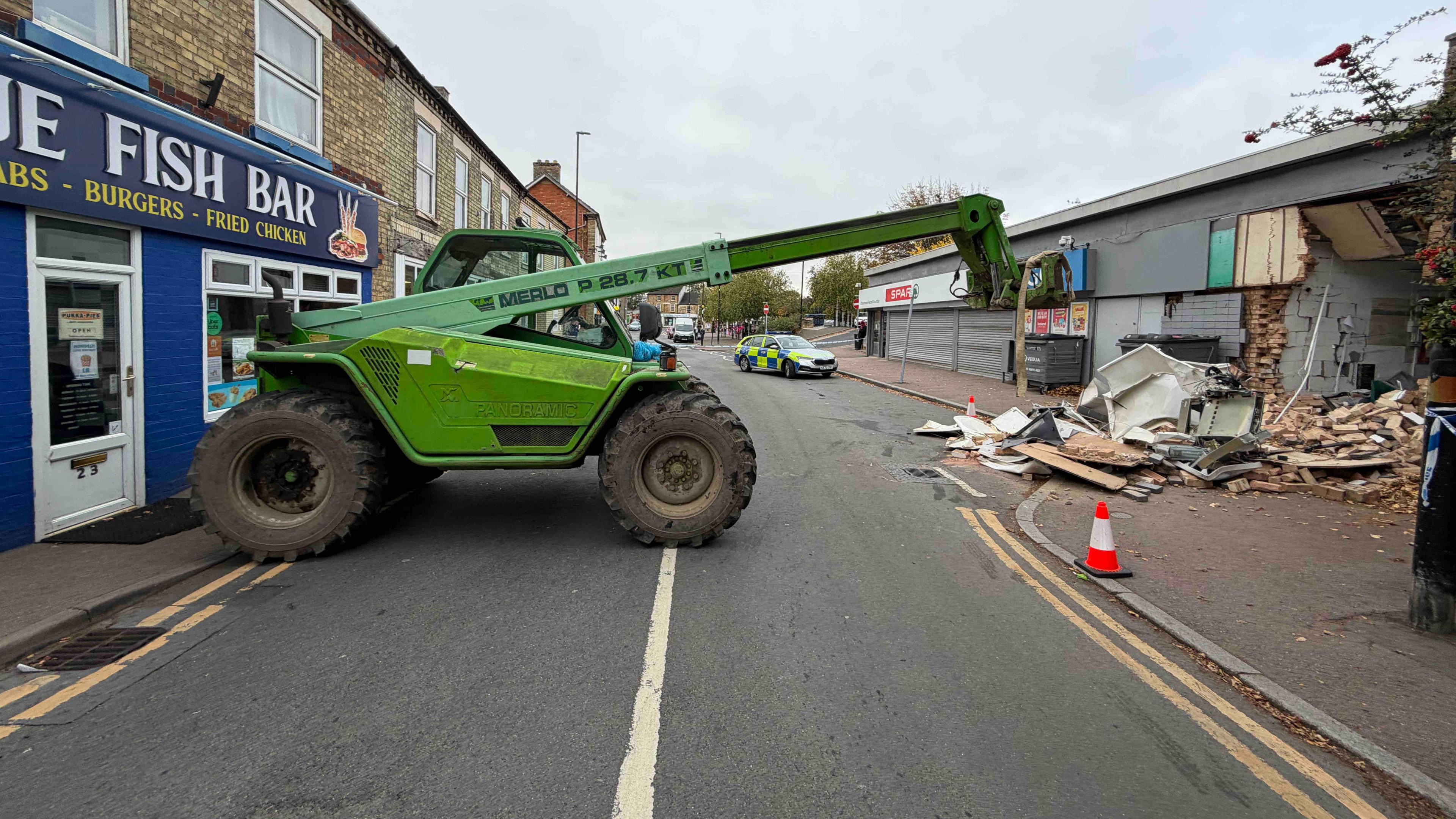 A telehandler forklift parked across a road. The damaged supermarket is roughly in line with the forklift mechanism. 
