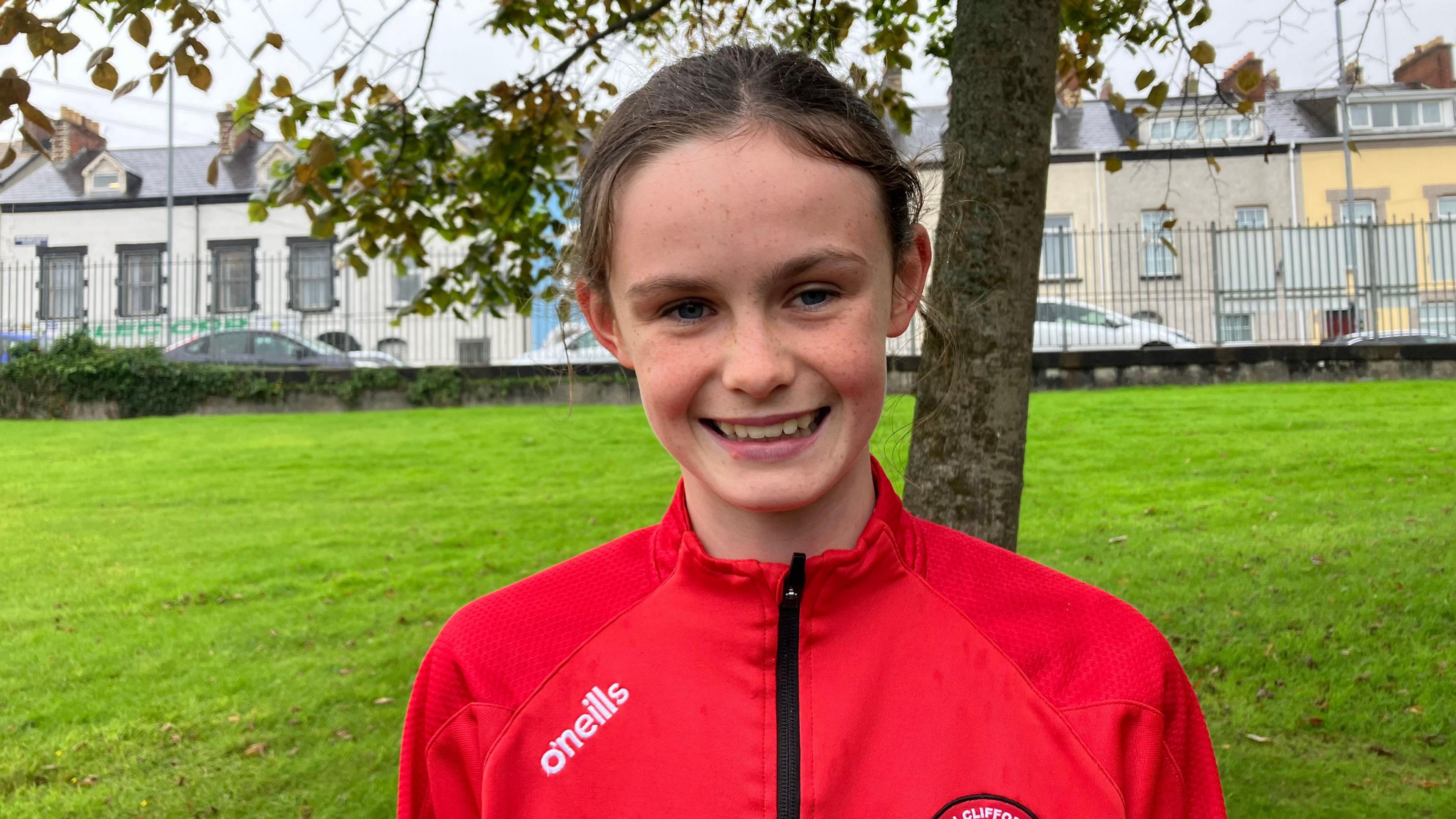 Zara. She has brown hair tied back, wearing a red zip-up football top.  She is standing in a field with a tree and houses in the background.