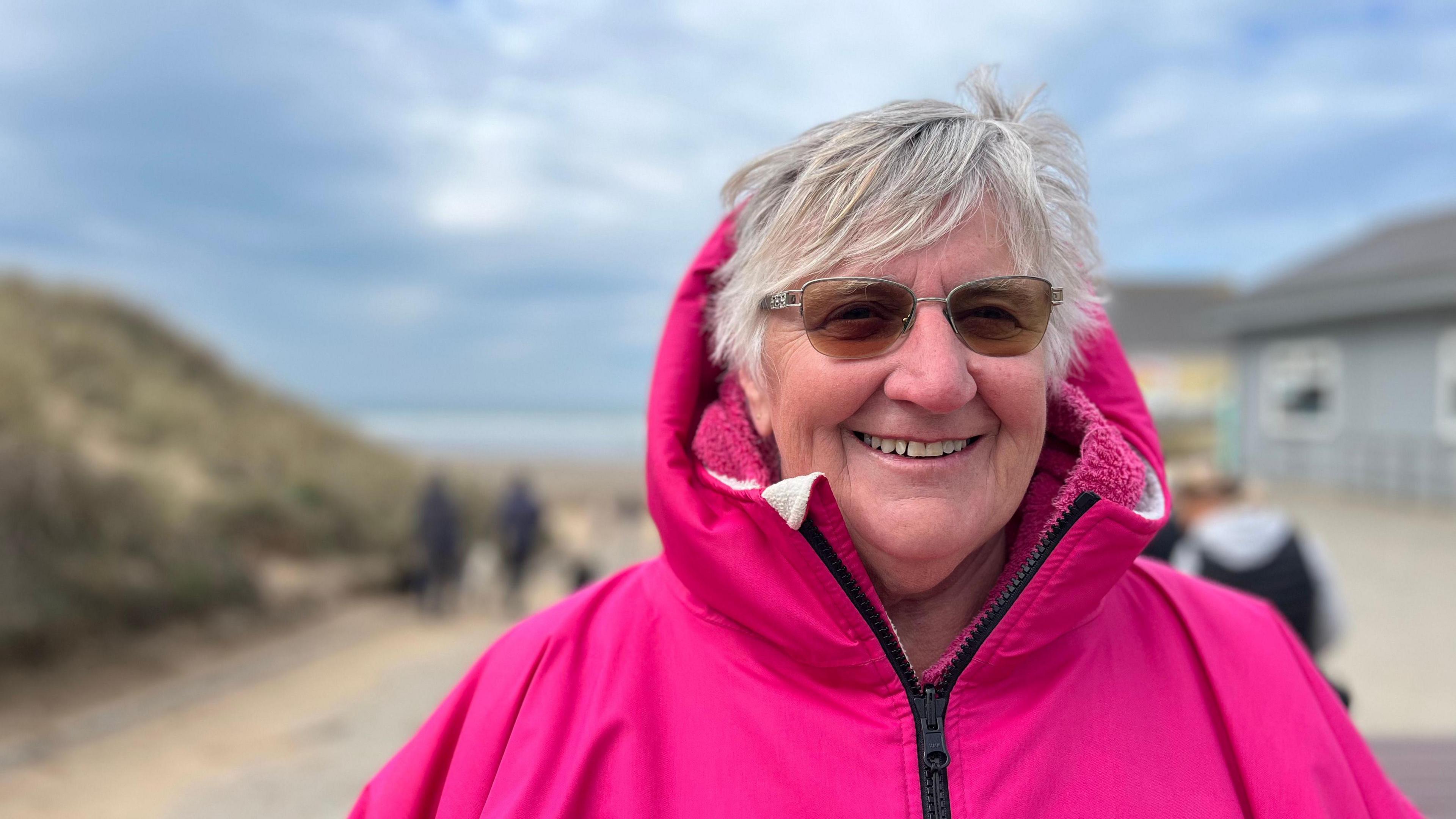 A picture of Lynda Price wearing a bright pink coat and glasses. She is stood in front of a path leading down to the beach.