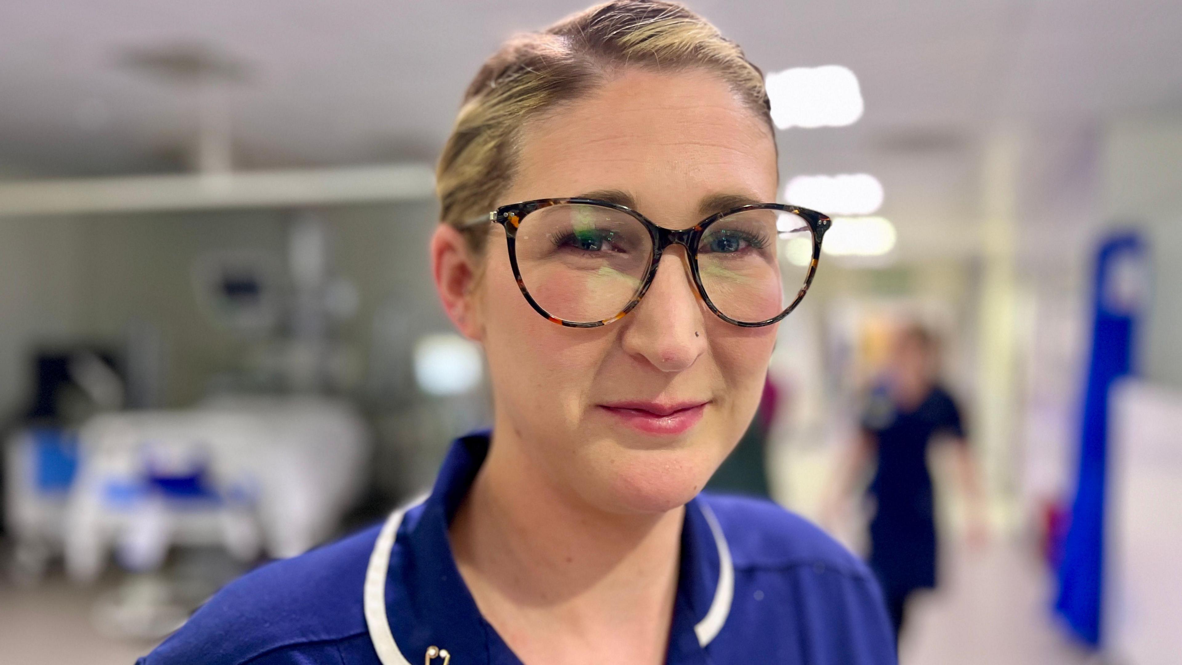 Senior sister Lauren Jakes stands in the critical care department at the Norfolk and Norwich University Hospital  The background is blurry but you can see a bed to the left of her and another nurse to the right. She is wearing a blue uniform with her hair plaited back and tortoiseshell glasses.