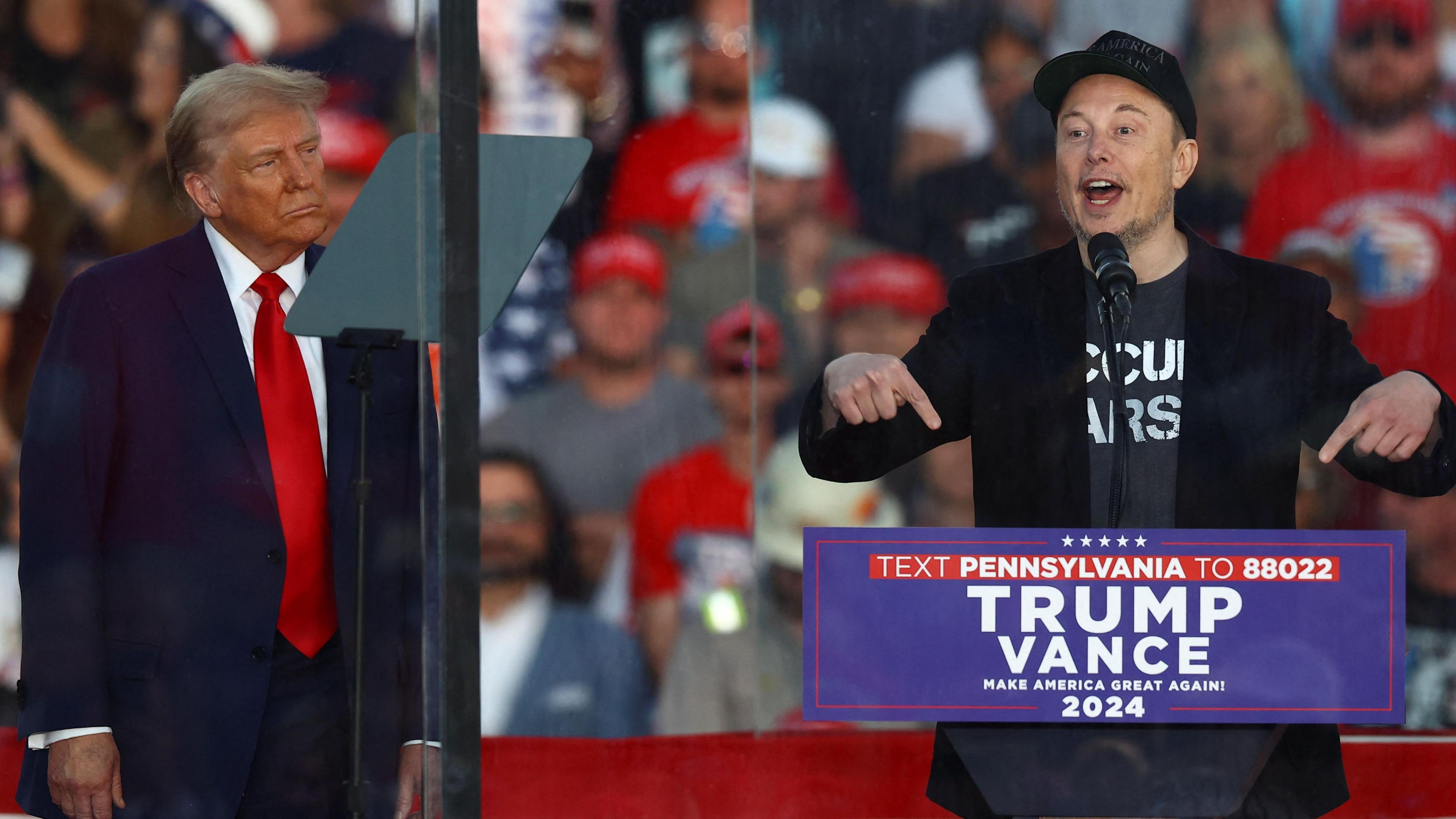 Donald Trump watches on stage as Musk, standing to his left, speaks at a podium reading 'Trump Vance Make America Great Again 2024' during a rally. Behind them is a blurry crowd