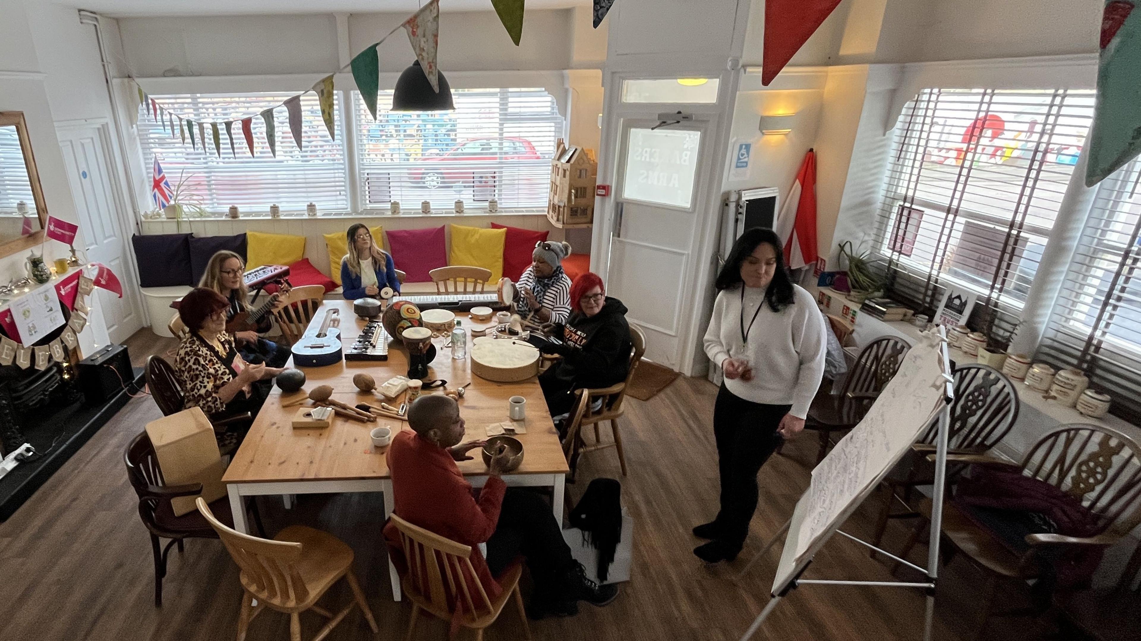 Six women sat around a table playing different instruments looking at a flip char for the song's words