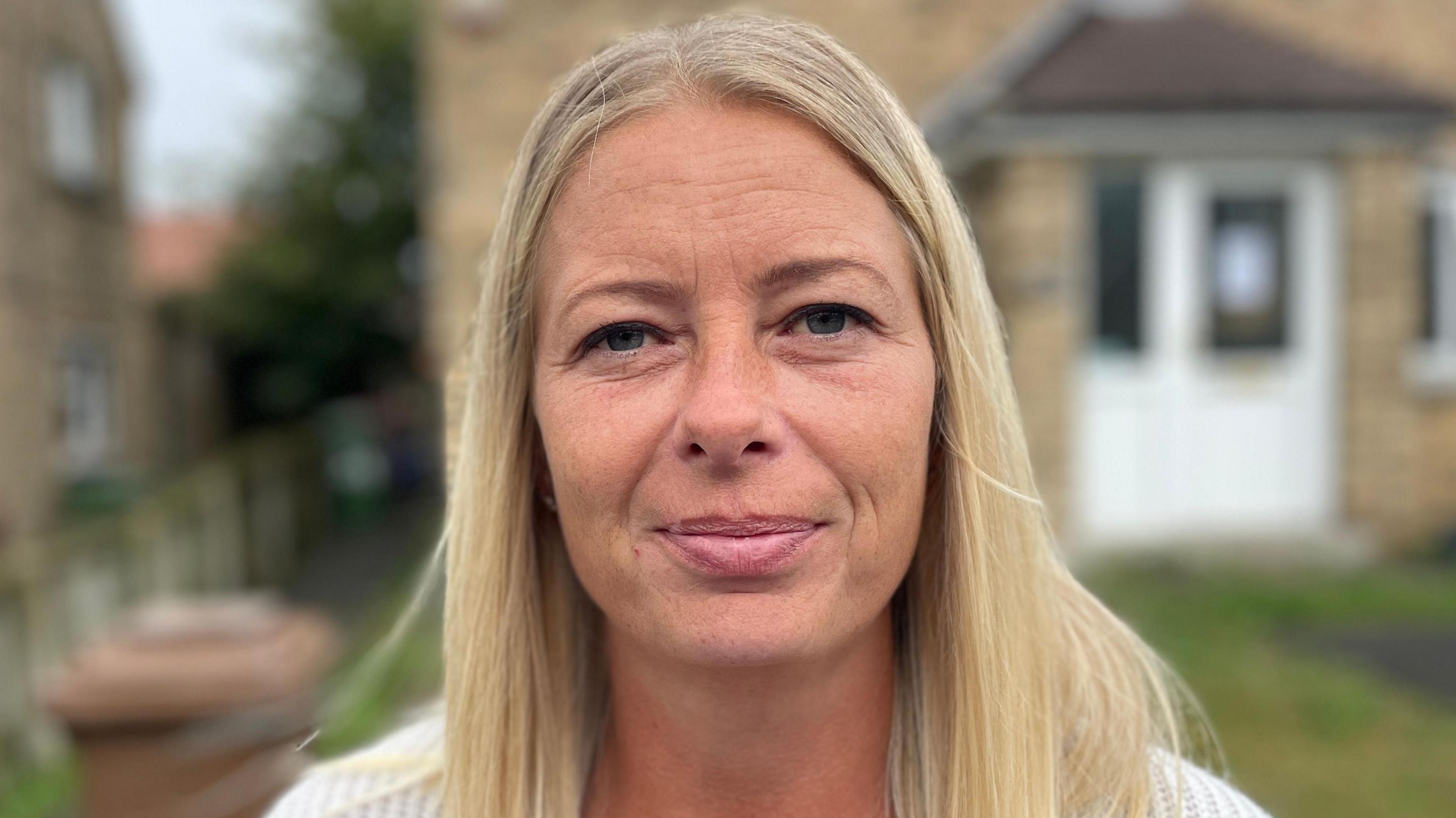 Donna Smethurst is smiling, she has long blond hair and blue eyes. She's wearing a white cardigan and is standing in front of a house with a white door. The background behind Donna is blurred. 