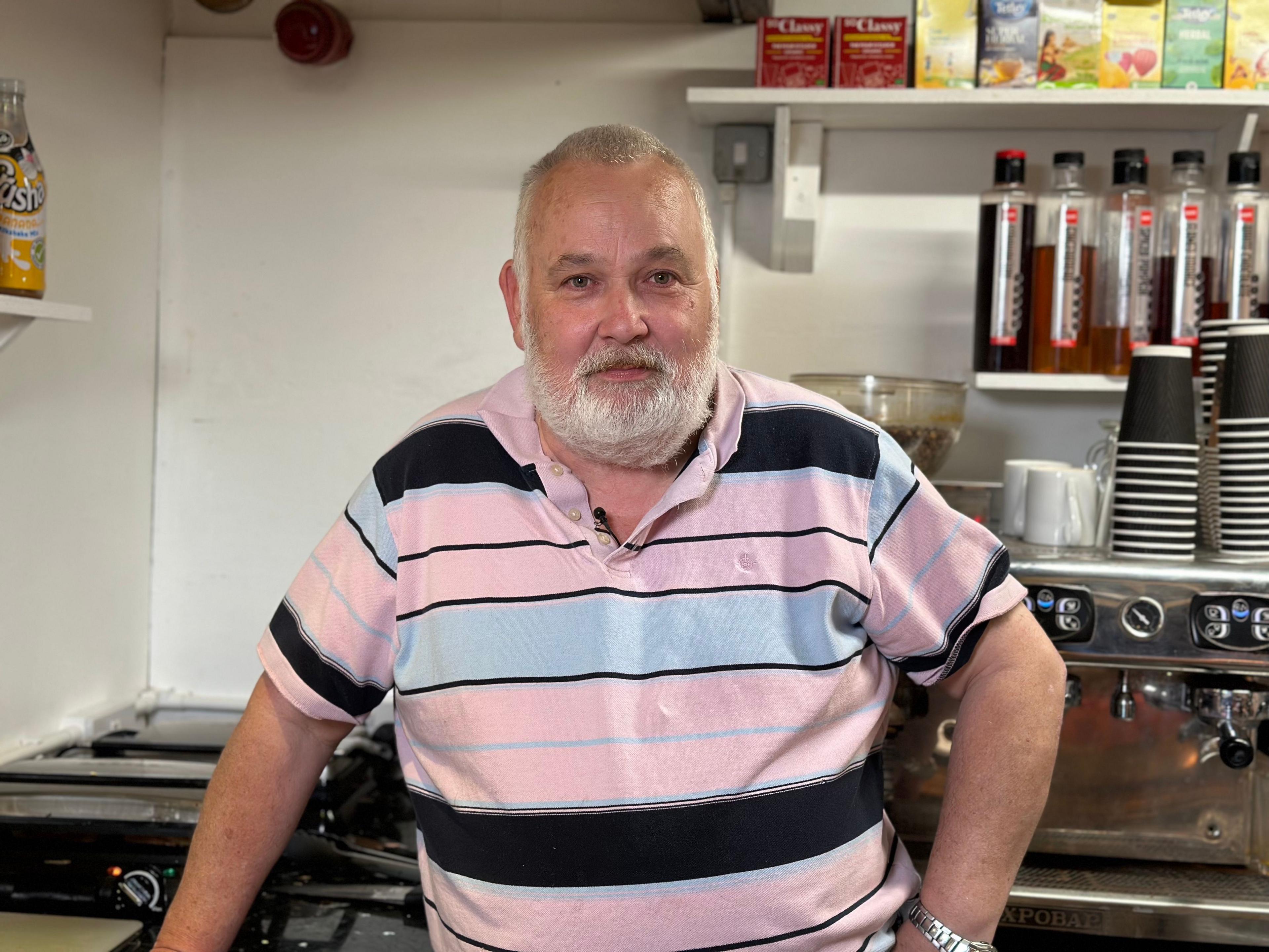 Mark Breen with a grey beard, wearing a pink striped polo shirt standing in his cafe kitchen with mugs and disposable cups seen behind him