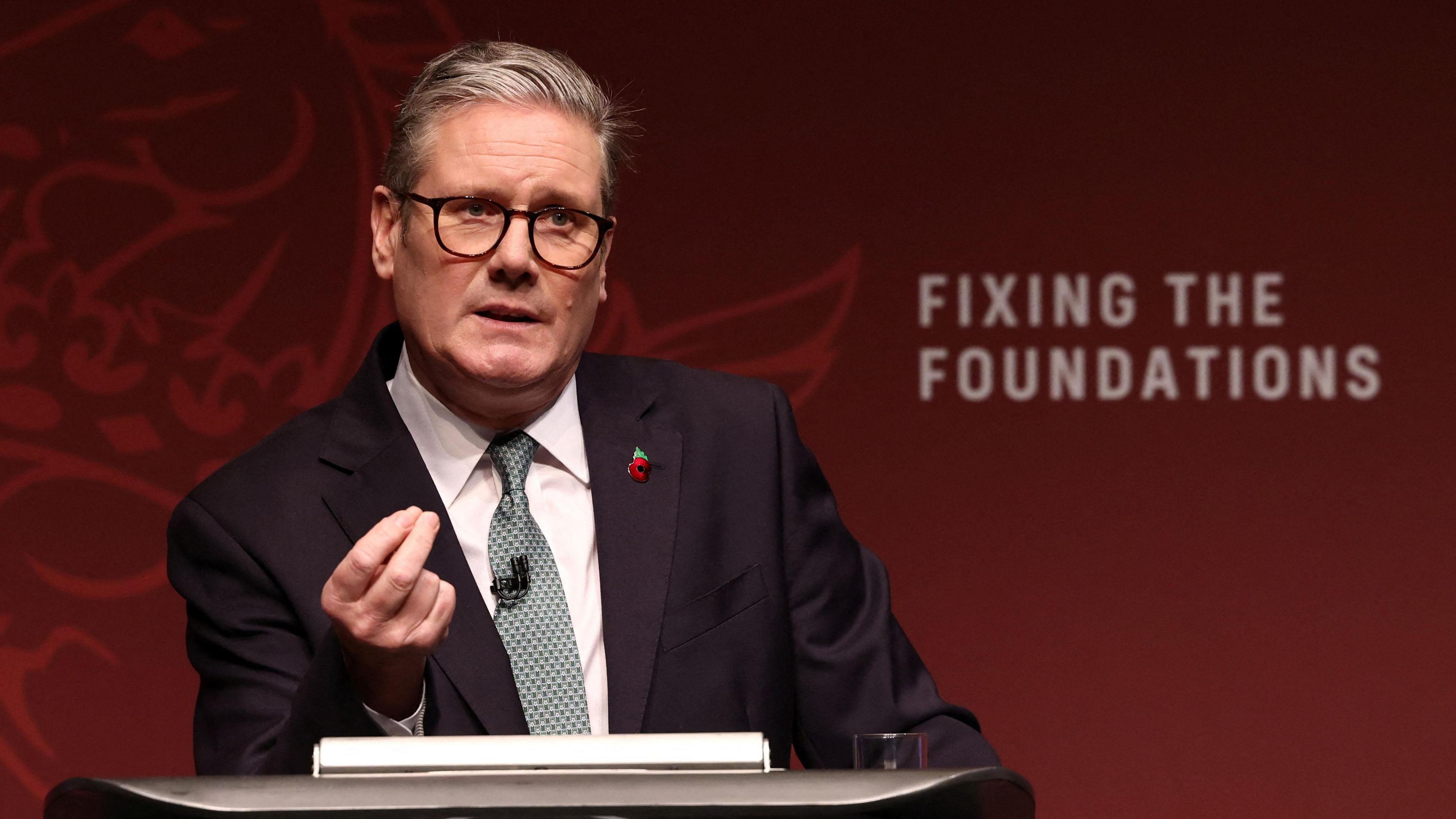 Prime Minister Sir Keir Starmer making a speech. He is wearing a suit and a poppy pin. He has a green and white tie and is making a point with his hands. He is standing in front of a wall that reads 'fixing the foundations'. 